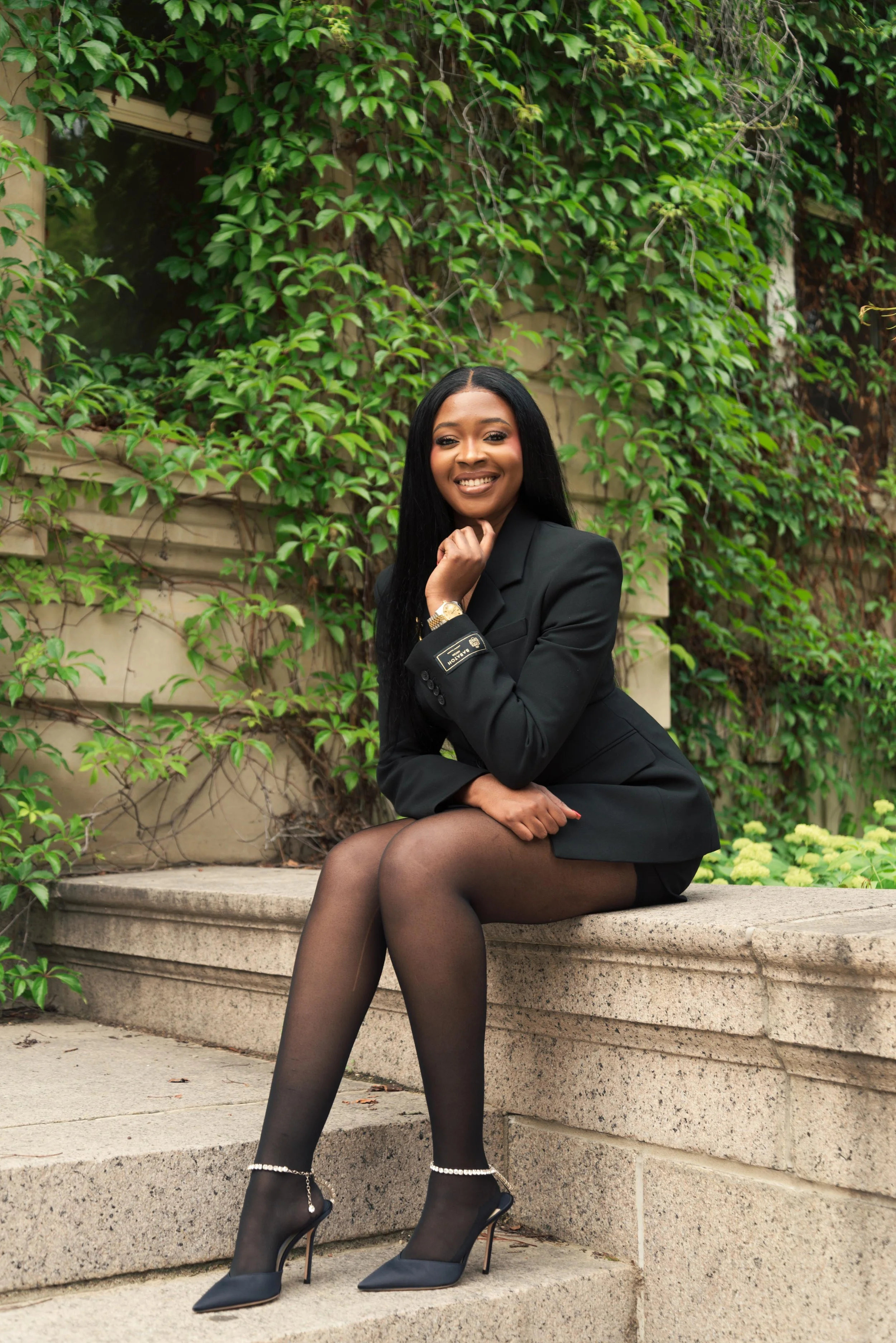 A woman sitting on a stone bench outdoors, dressed in a black blazer, black stockings, and black high heels with ankle straps, smiling at the camera, with green vines growing on a wall behind her.