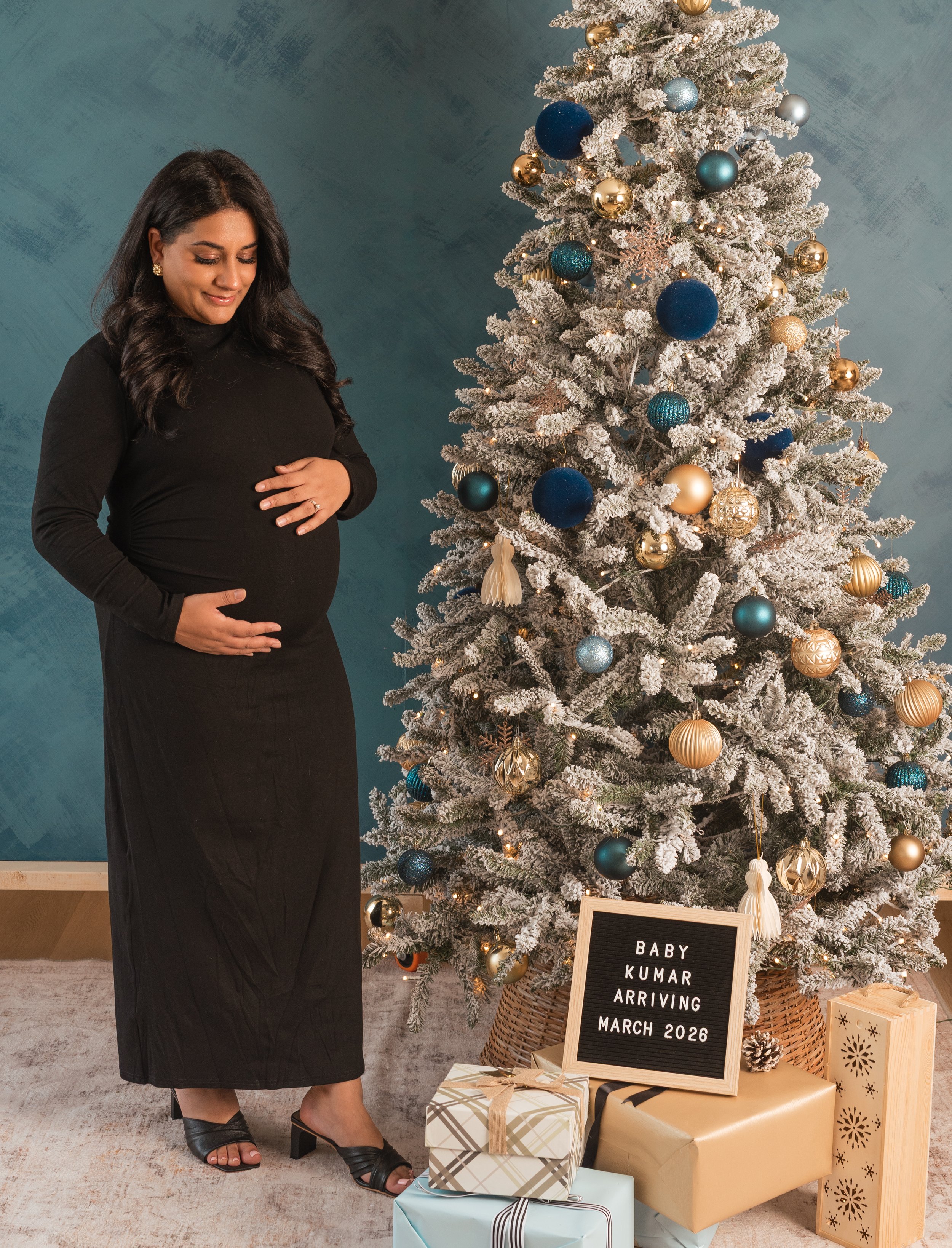 A pregnant woman with dark hair wearing a black dress and black heels stands beside a decorated Christmas tree. The tree is adorned with gold, blue, and silver ornaments, and there are wrapped gifts beneath it. A signboard reads, 'Baby Kumar arriving