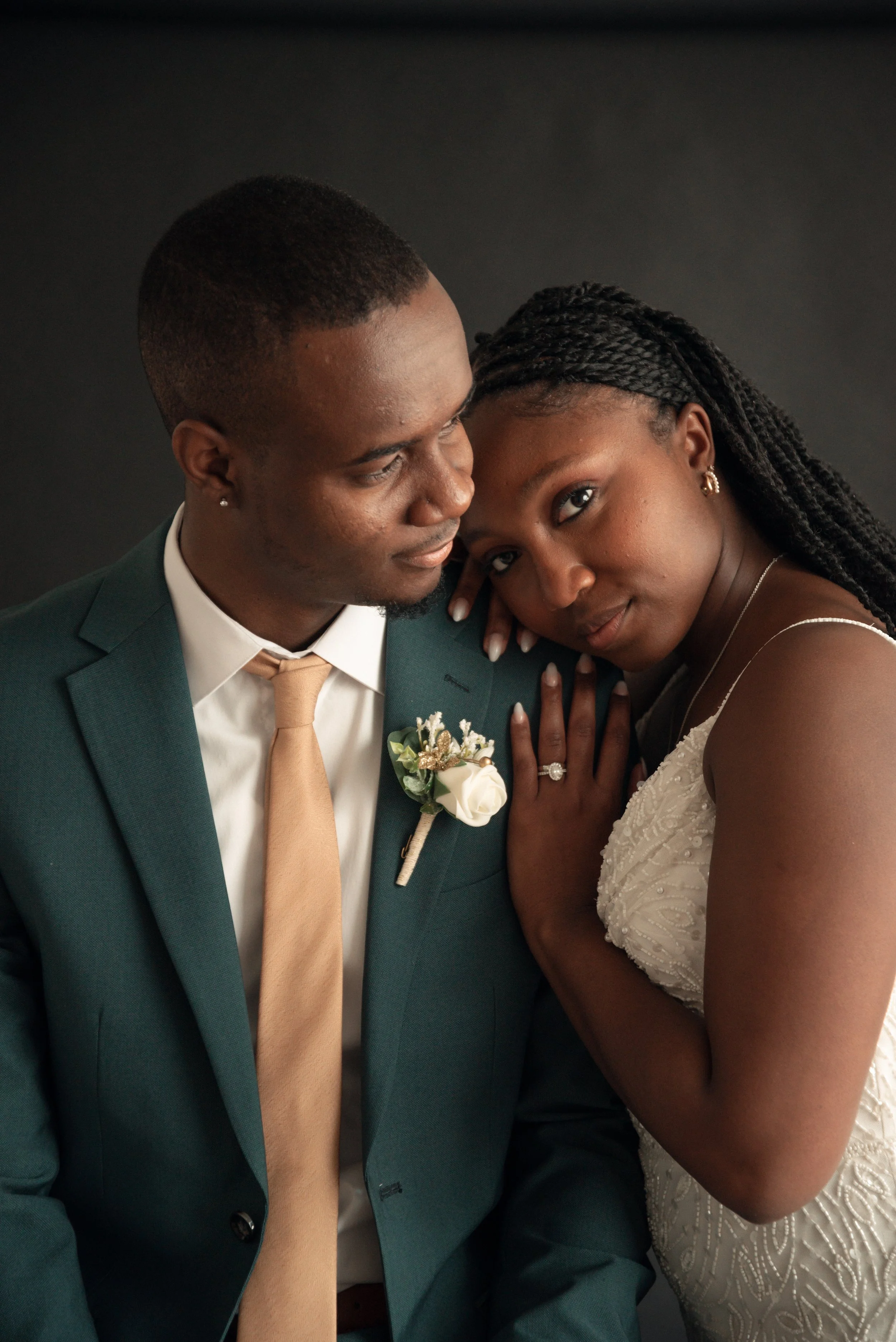 A close-up of a bride and groom in wedding attire, with the bride resting her head on the groom's shoulder and both looking at each other affectionately against a dark background.
