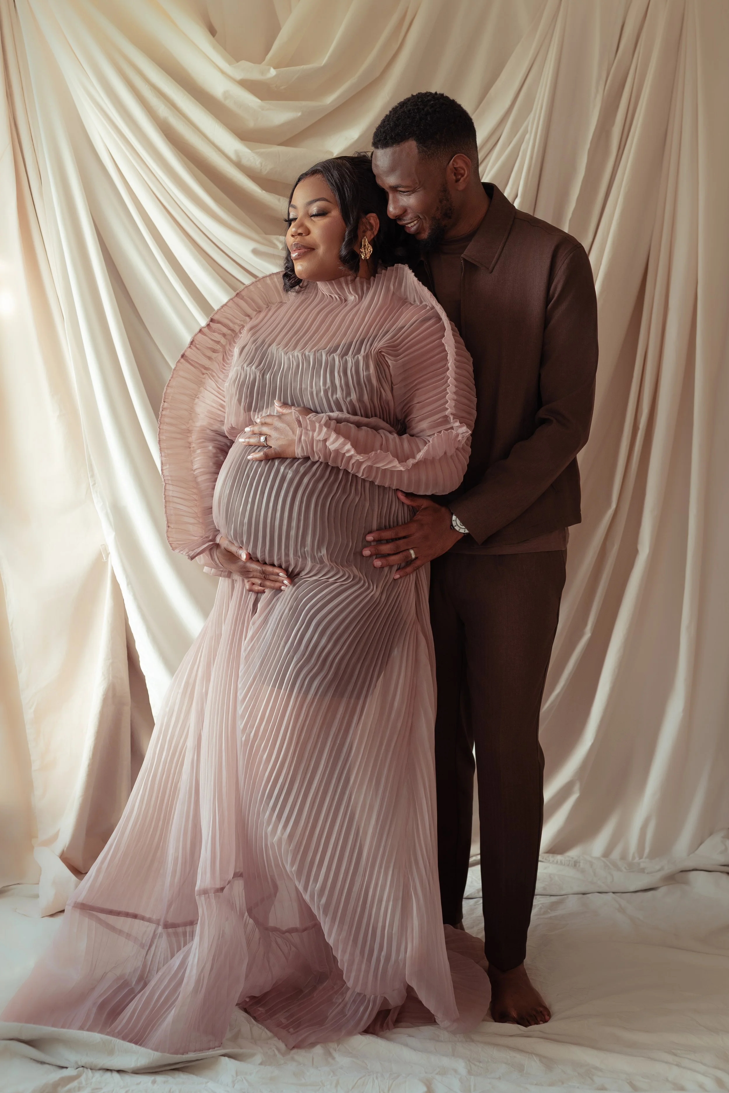 A pregnant woman in a flowing, pleated pink dress stands with her eyes closed, resting her hand on her belly, while a man behind her gently holds her waist. They are smiling softly in front of a cream-colored curtain backdrop.