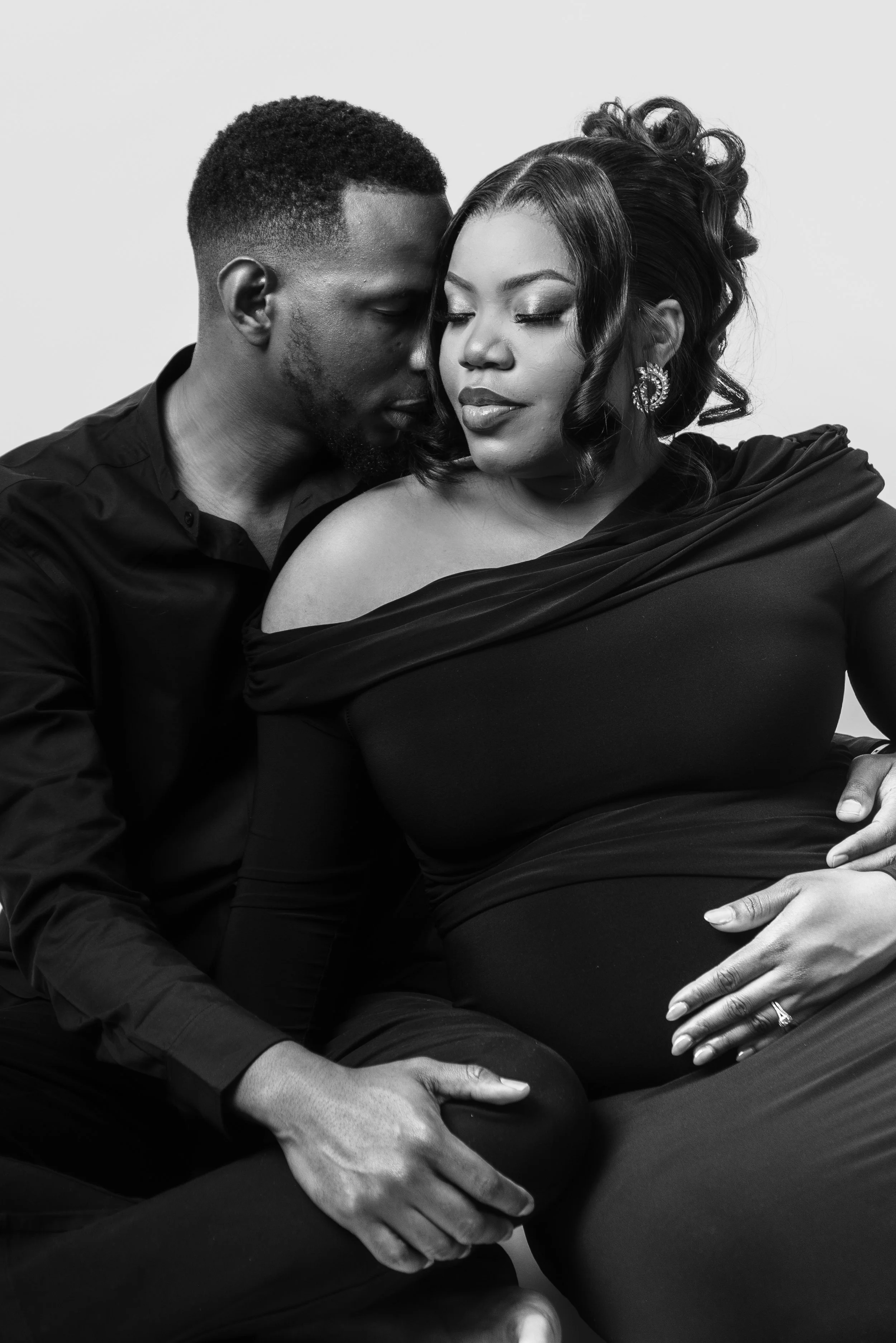 A black and white photograph of a man and a pregnant woman in an intimate pose, with their eyes closed and foreheads touching. The man is sitting behind the woman, gently holding her belly, and the woman is wearing earrings and a dress.