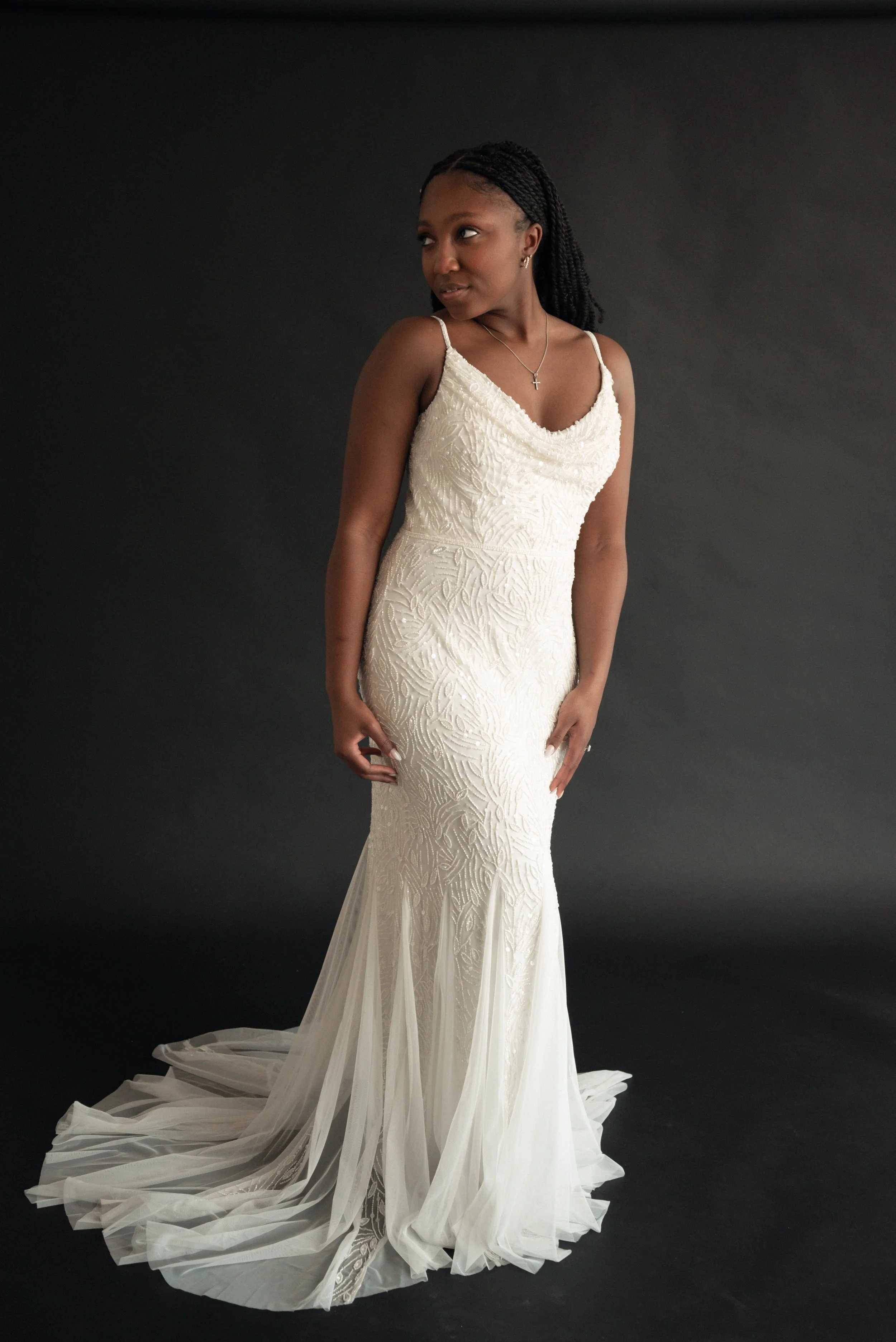 A woman in a white wedding dress with intricate embroidery, standing against a black background, with a long train flowing on the floor.