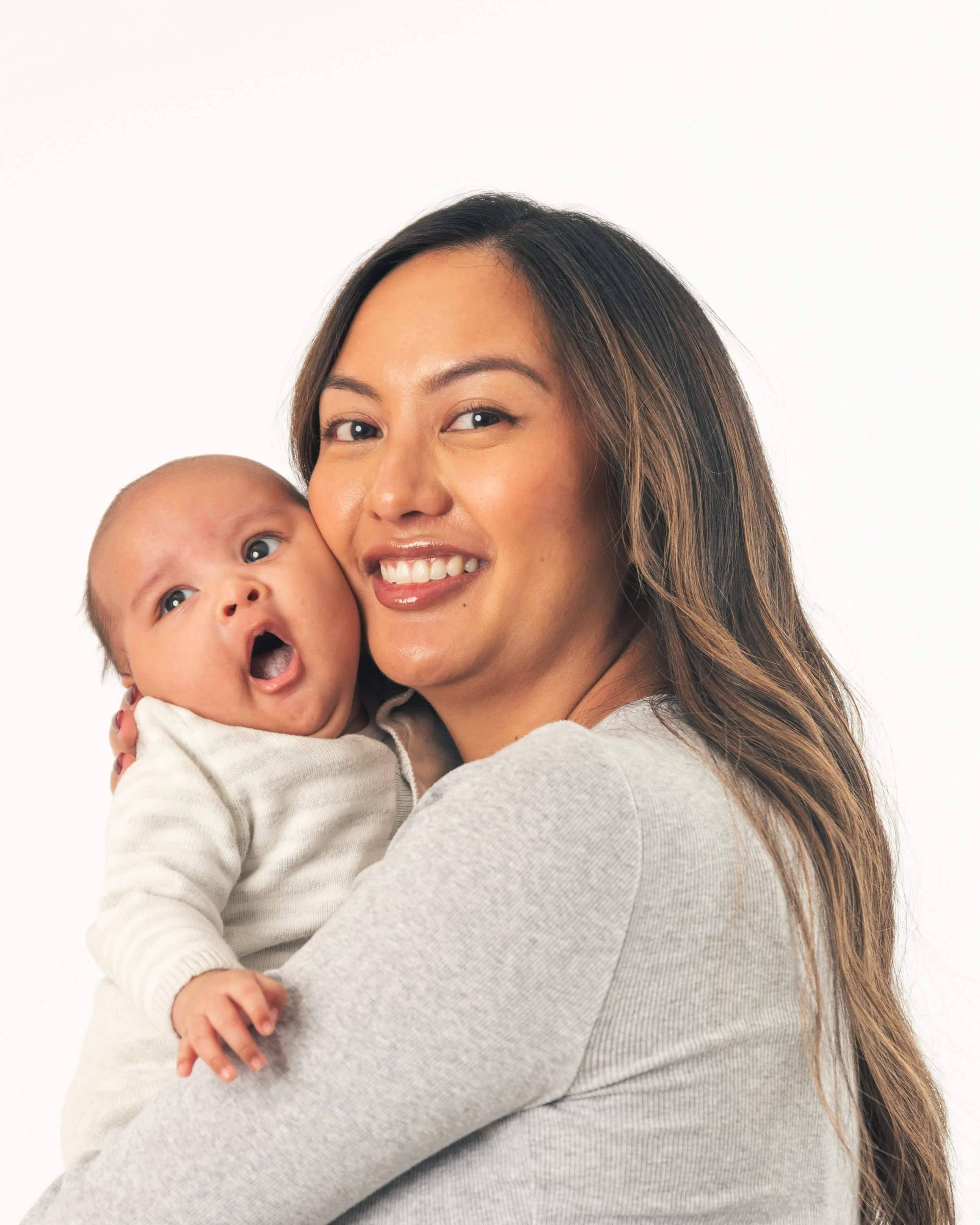 A woman with long, wavy hair smiling and holding a baby close to her face against a plain white background.