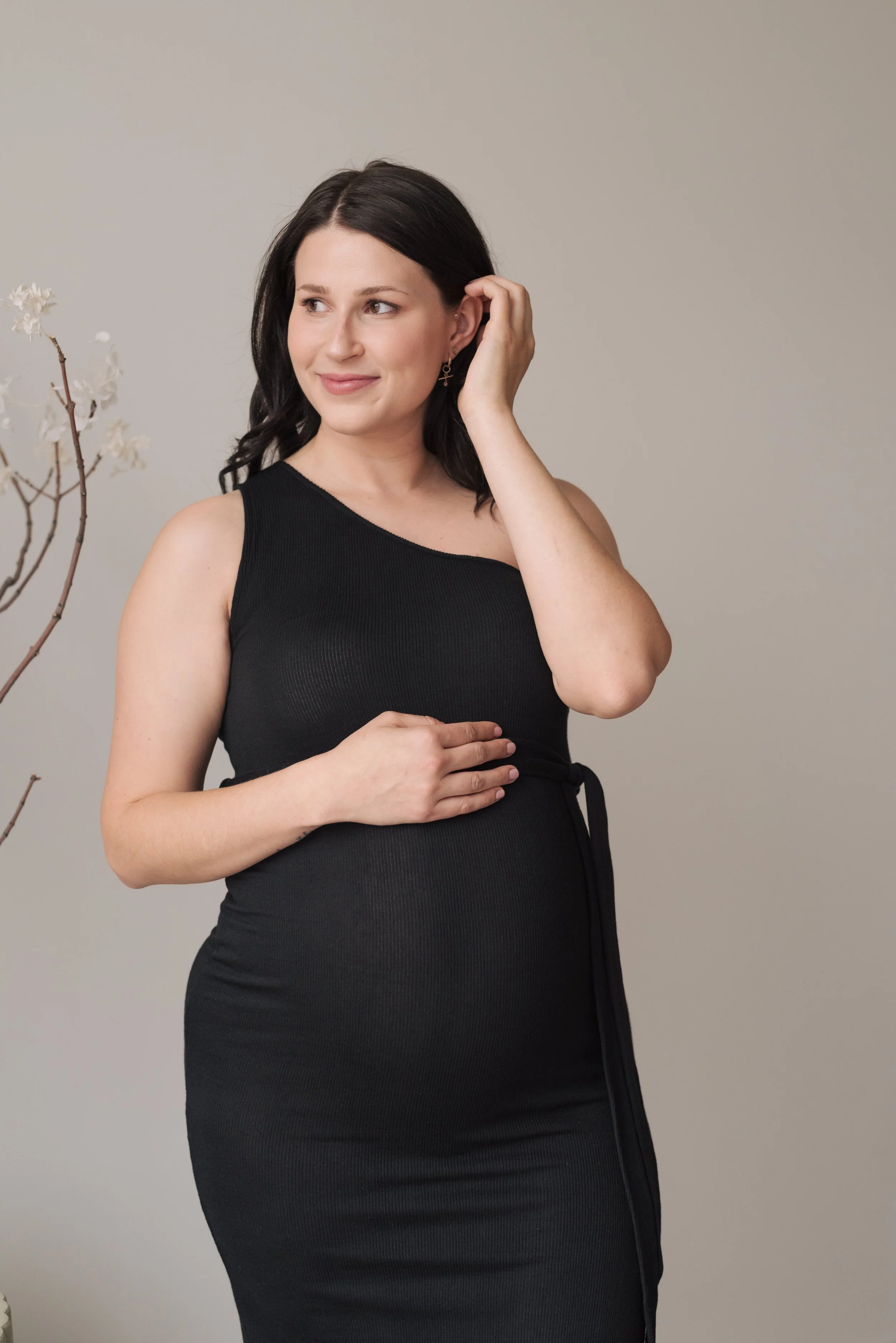A woman with dark hair wearing a black sleeveless dress, touching her earring with her right hand and resting her left hand on her belly, standing next to a flowering branch.
