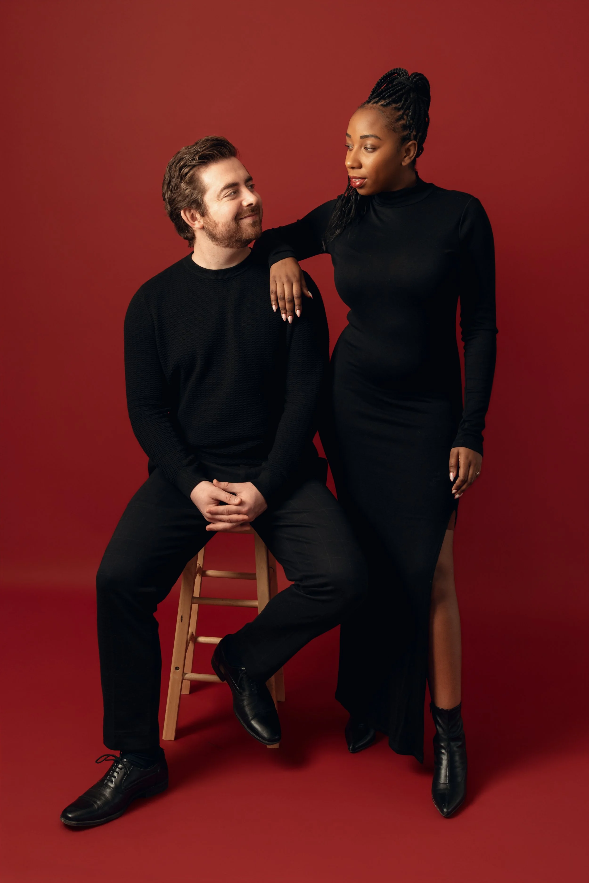 A man and a woman with dark skin posing together against a red background. The man is sitting on a wooden stool, wearing a black sweater and pants, with black shoes. The woman is standing with one arm resting on the man’s shoulder, wearing a black dr