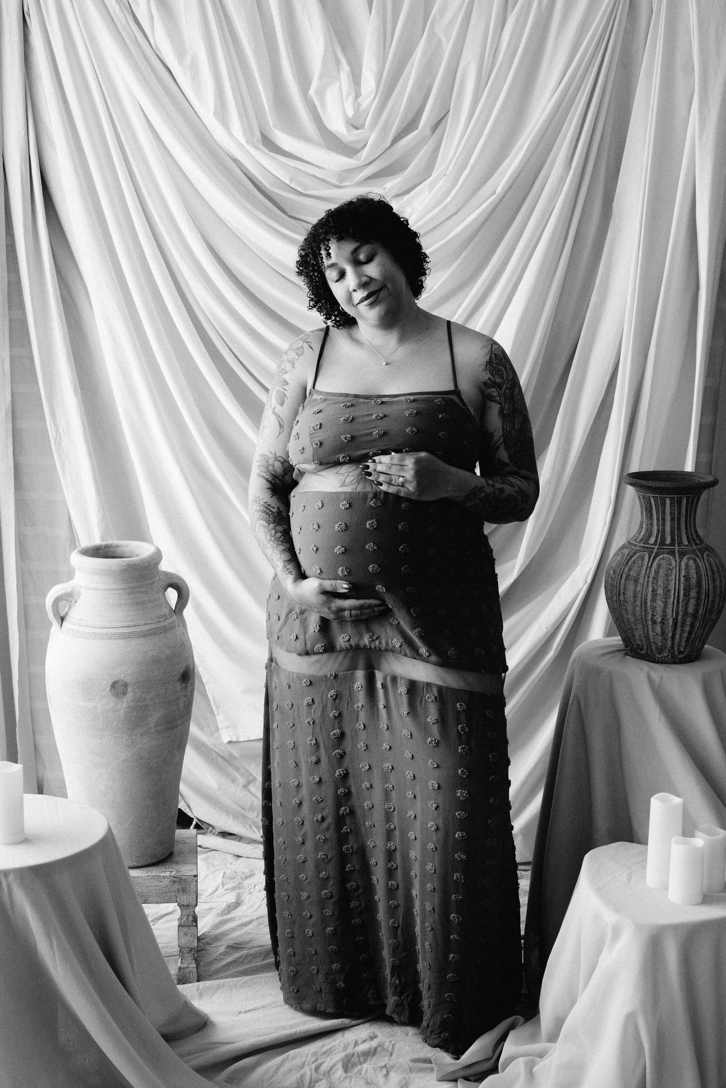 Black and white photo of a pregnant woman standing with eyes closed and a peaceful expression. She is wearing a spaghetti strap dress with floral embroidery and has tattoos on her arms. She is holding her belly with one hand. The setting includes dra