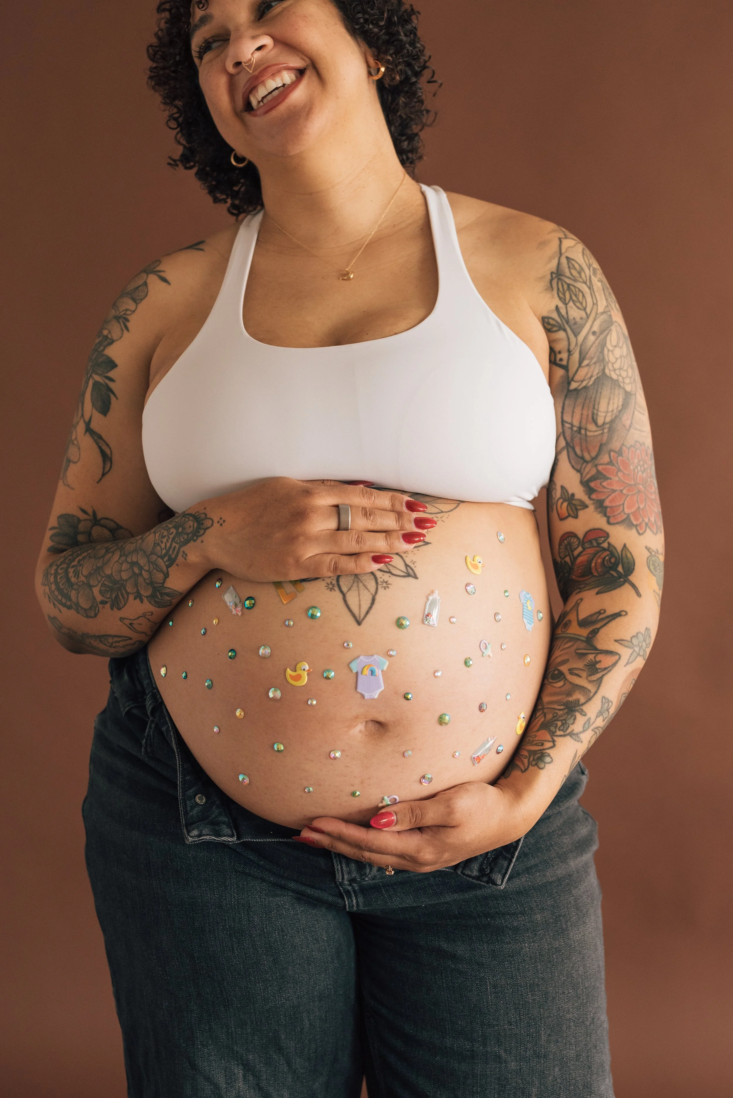 A woman with curly dark hair, tattoos, and multiple piercings wearing a white sports bra and jeans, smiling and holding her pregnant belly which is decorated with colorful stickers, against a brown background.