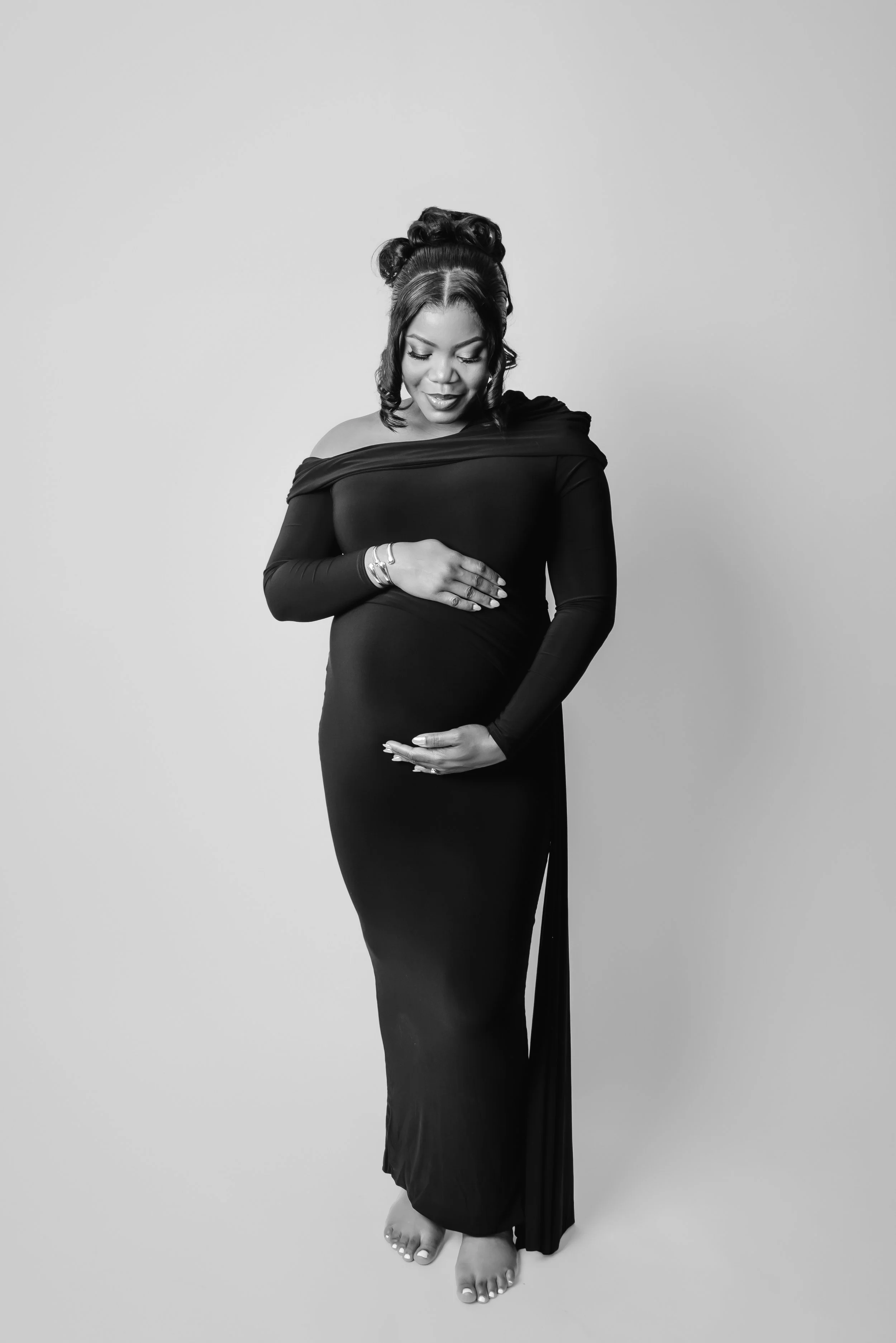 Black and white photo of a woman standing barefoot, wearing a long, fitted dress, with her eyes closed, smiling softly, holding her belly with both hands, and her hair styled in an elegant updo.