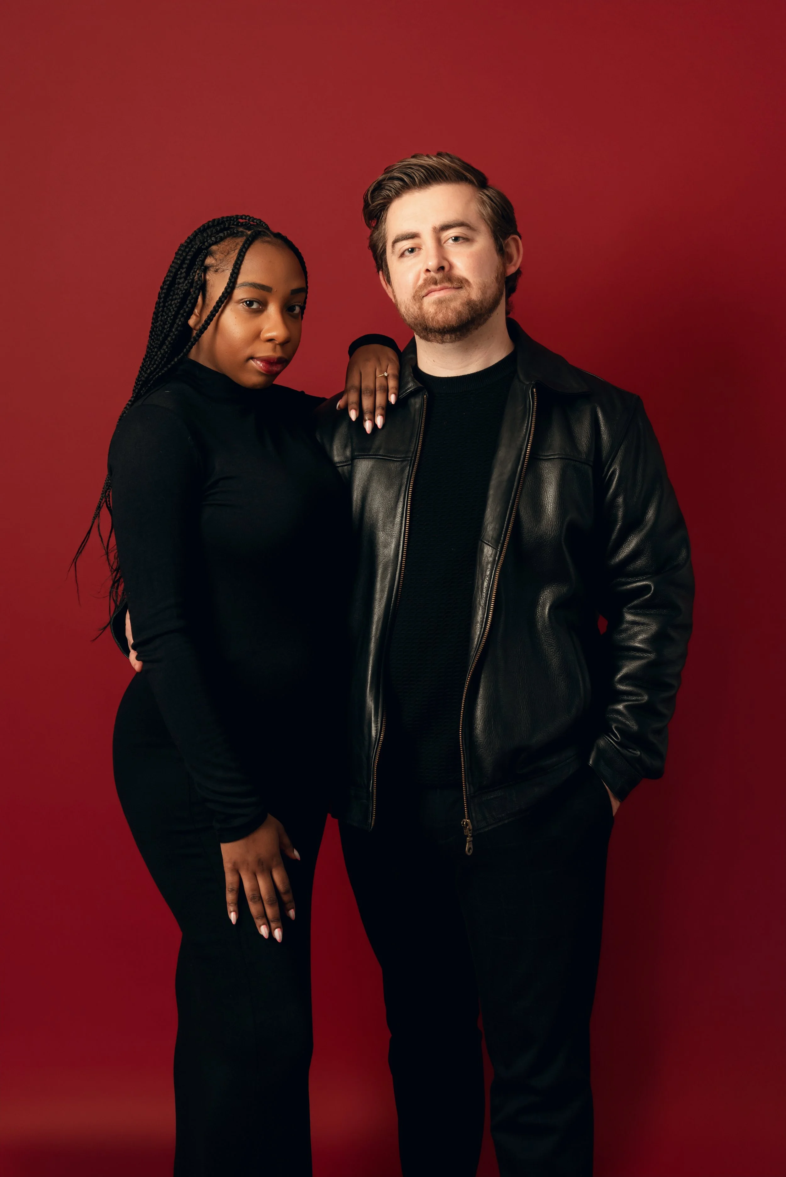 A young woman and a young man stand against a red background. The woman has long braided hair and is wearing a black long-sleeve top and black pants. The man has short hair and facial hair, and is wearing a black leather jacket and black pants. The w