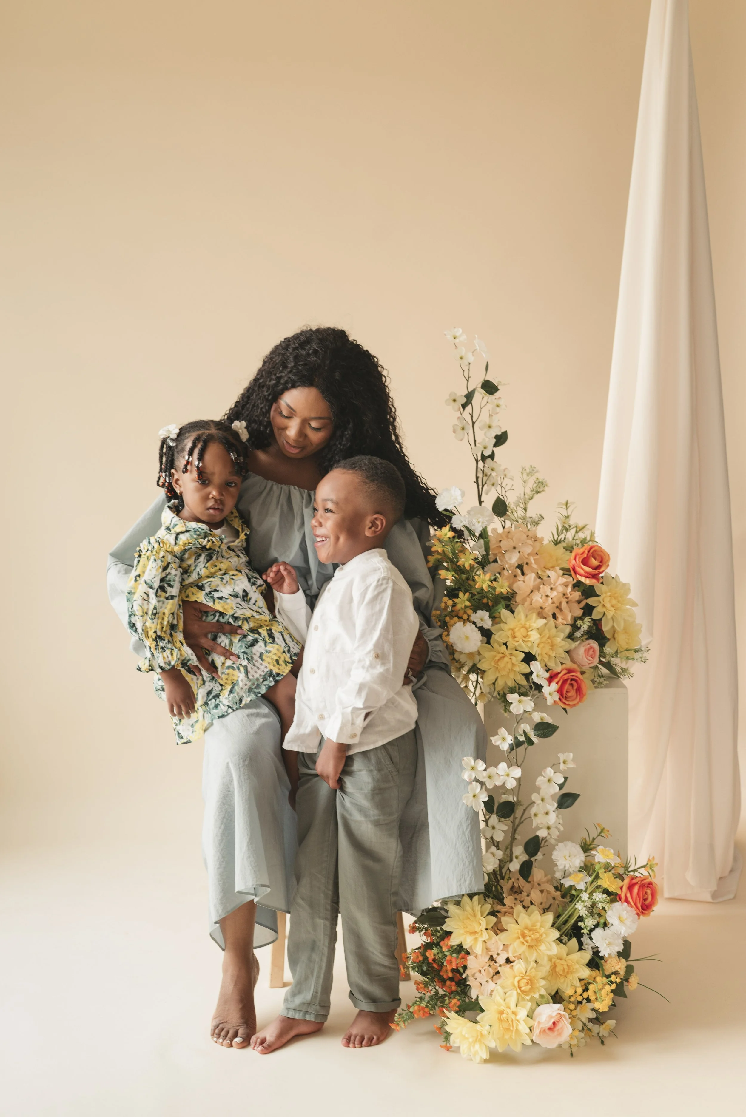 A woman with dark curly hair holding a young girl with braided hair and flowers in her hair, along with a smiling young boy, in front of a large floral arrangement with yellow, white, and orange flowers against a beige background.