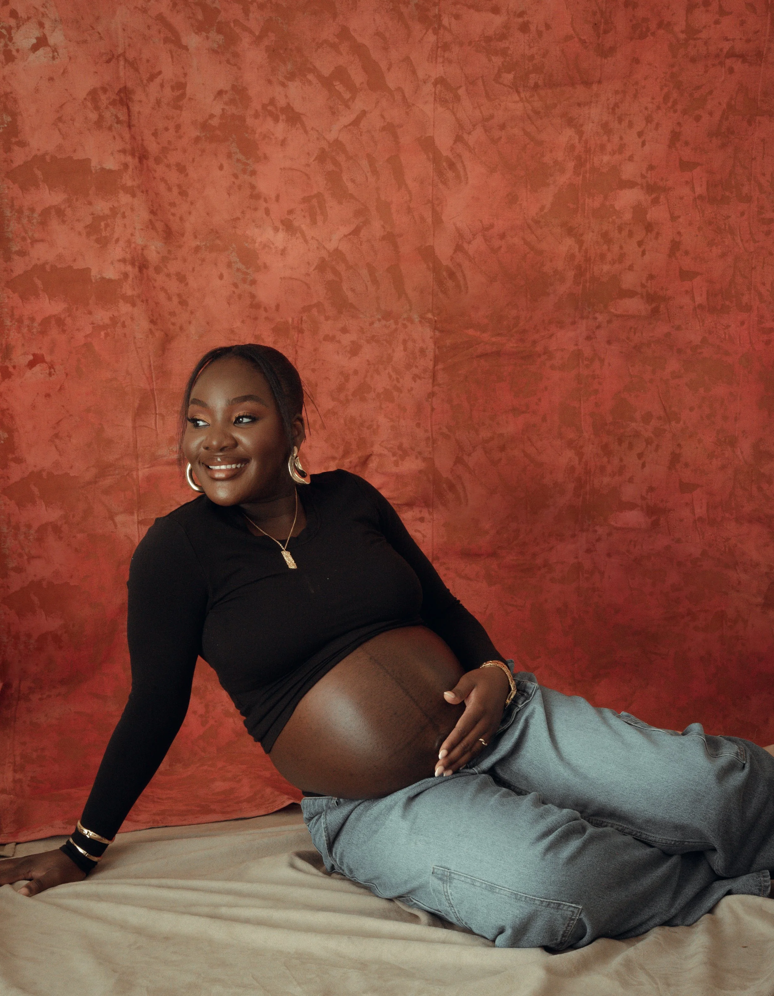 Young woman with a big smile, wearing black top, jewelry, and hoop earrings, sitting on a bed with red textured wall background.
