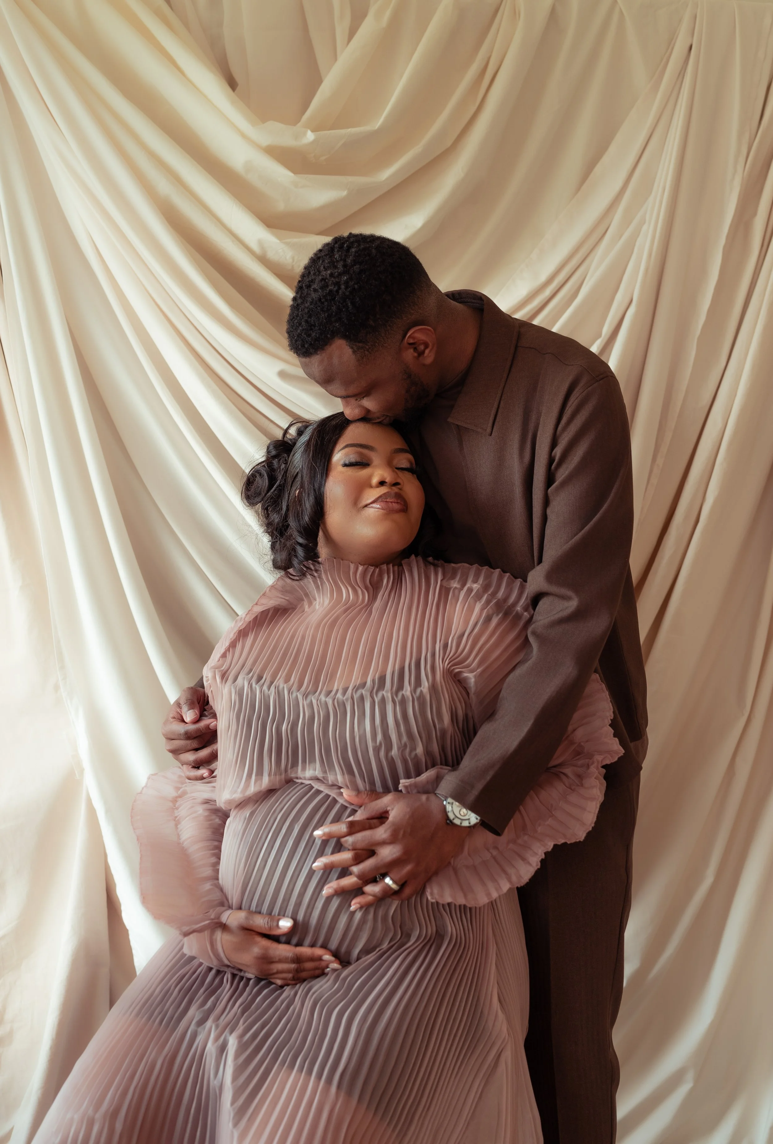A man gently kisses a woman on her forehead as she is held in his arms, with a cream-colored draped fabric backdrop.