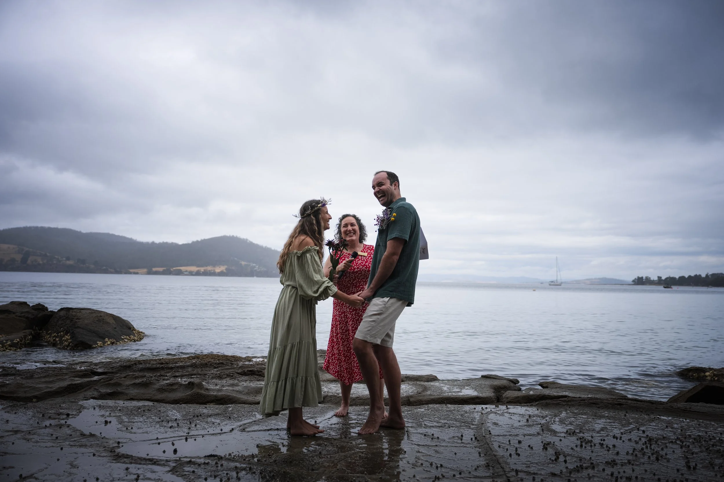 A couple standing barefoot on a stunning rocky beach in Tasmania holding hands and smiling at each other, with a woman officiating their wedding. The background features a beautiful beach in Bruny, hills in the distance, and a cloudy sky.