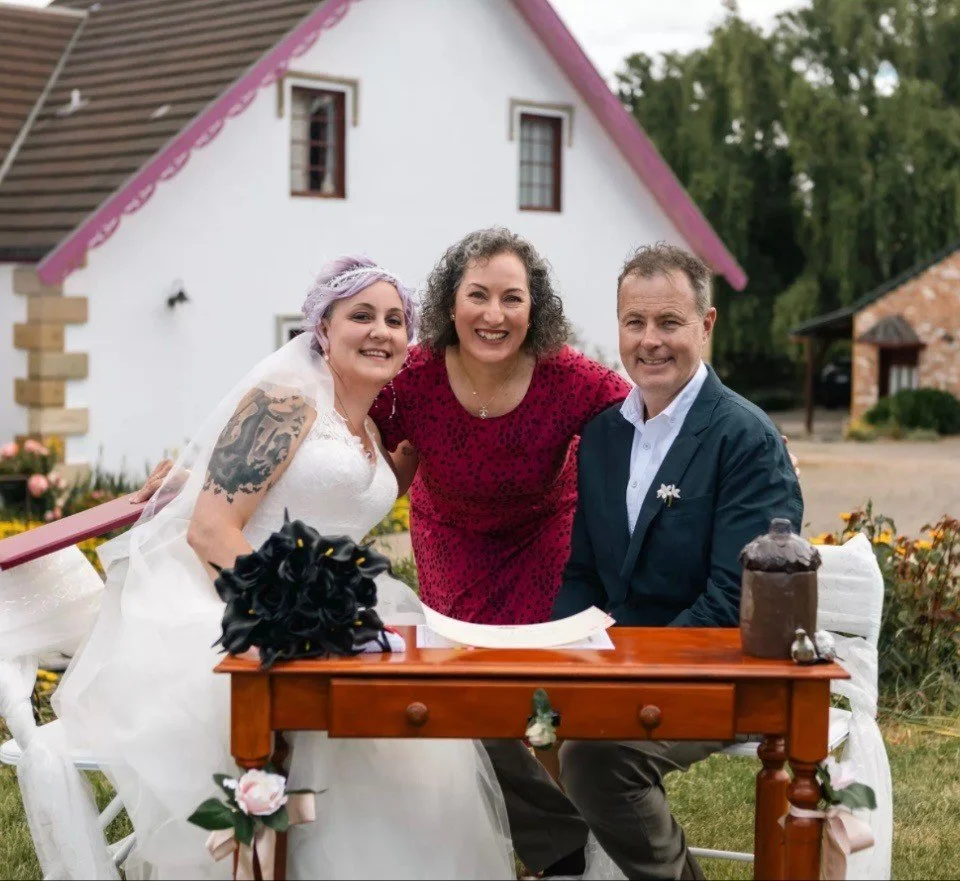 Beautiful bride and groom happy with their marriage celebrant smiling and posing together outdoors in front of a white cottage during a wedding celebration.