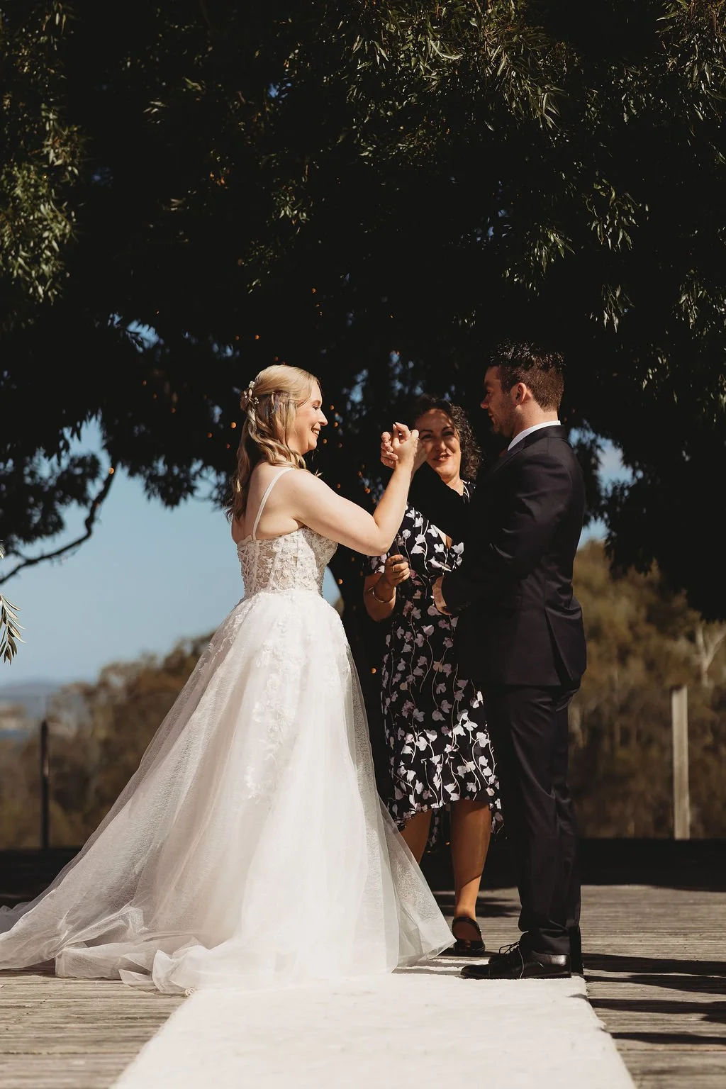 A bride and groom exchanging vows outdoors during their wedding ceremony, with an officiant standing behind them under a tree.