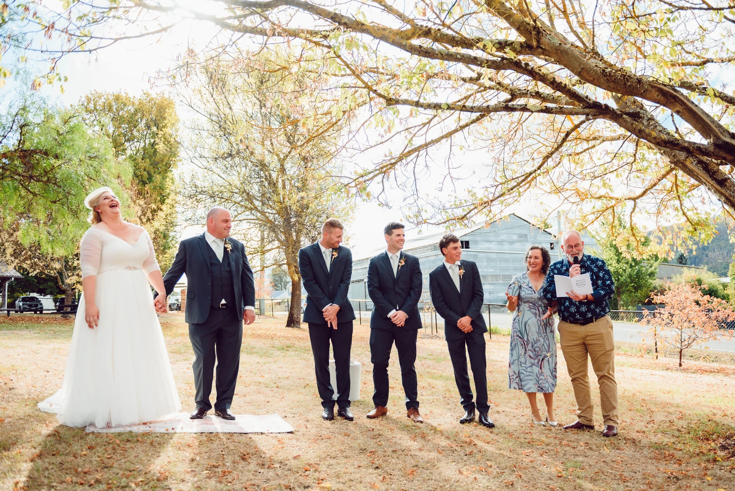 Bride and groom with wedding party standing outdoors under trees with autumn leaves, officiant reading from a paper.