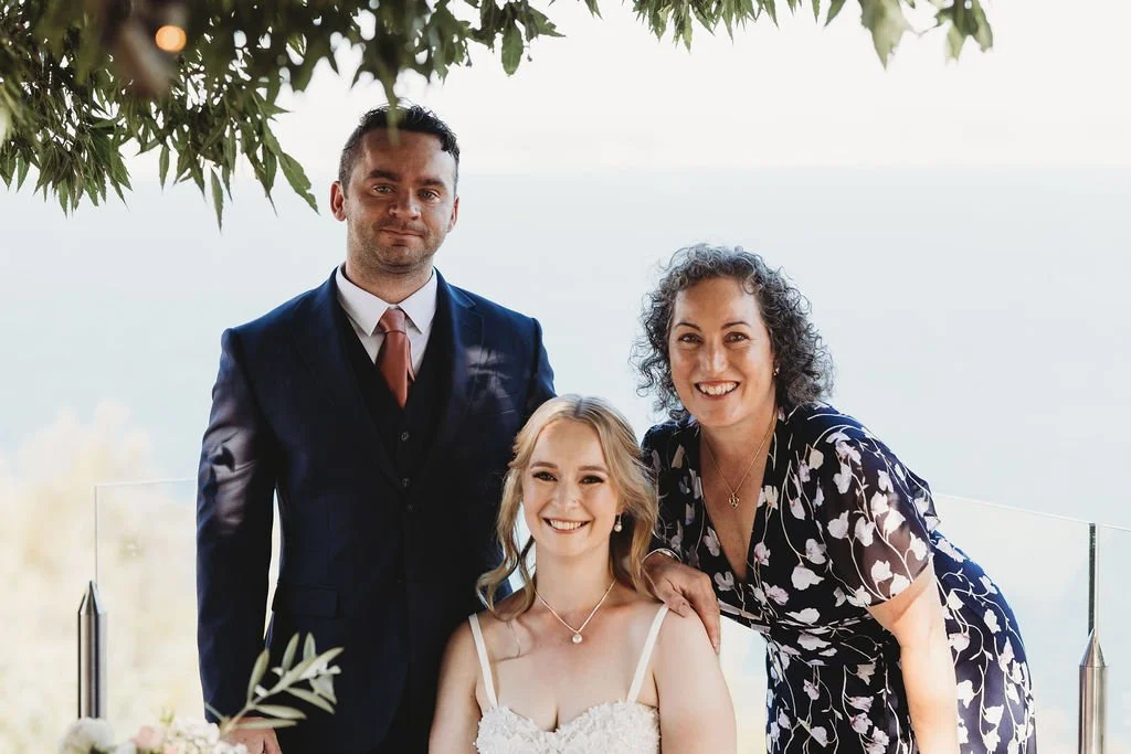 Three people at a wedding or formal event outdoors, with a woman in a white dress seated in the center, a woman in a floral dress standing on her right, and a man in a suit standing on her left, near a glass railing with a view of water.