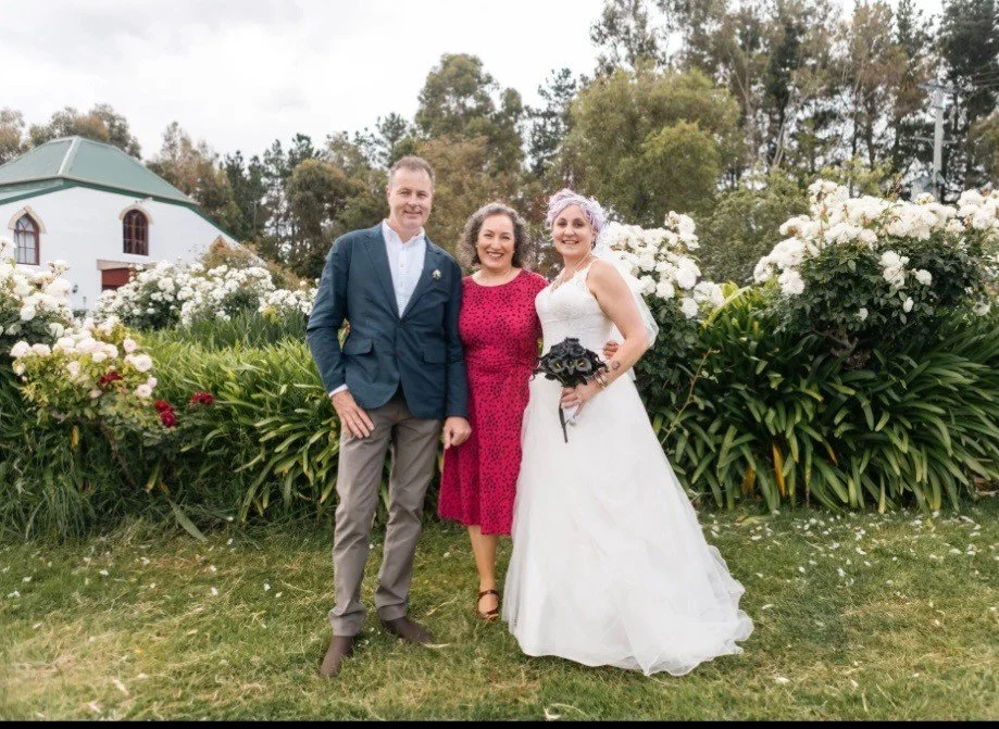 Bride and groom happy with their marriage celebrant in the outdoors in front of a garden of white flowers and green bushes- just after the wedding.