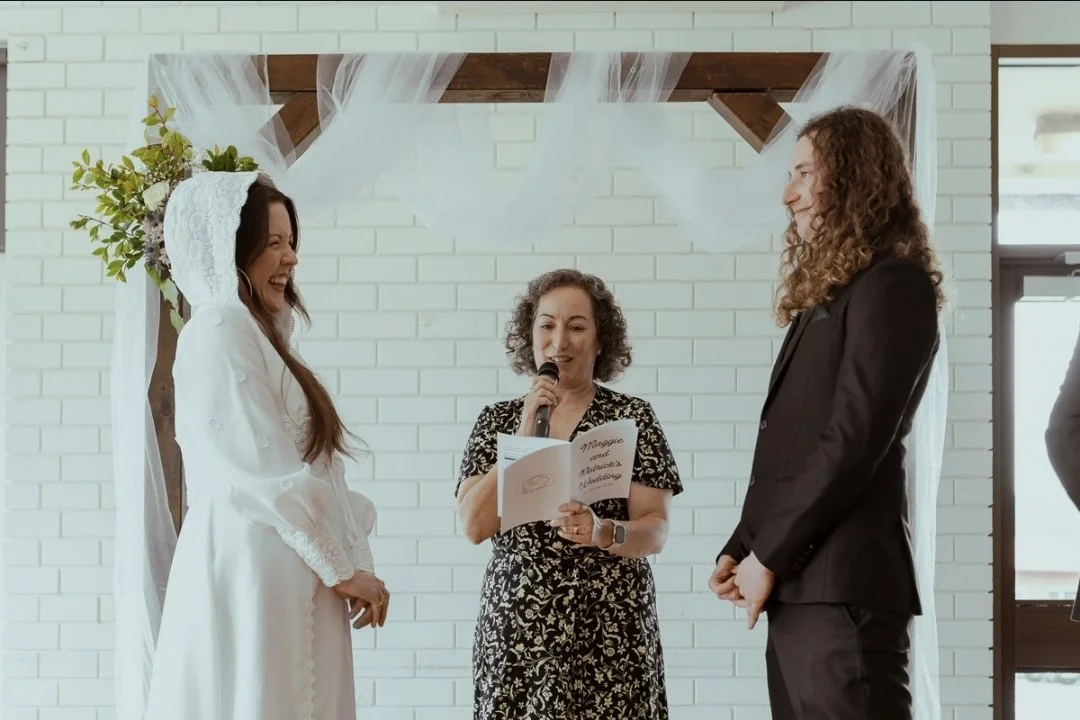 A couple standing during their wedding ceremony, facing each other and smiling, with a marriage celebrant holding her script and smiling with the couple at a community hall.