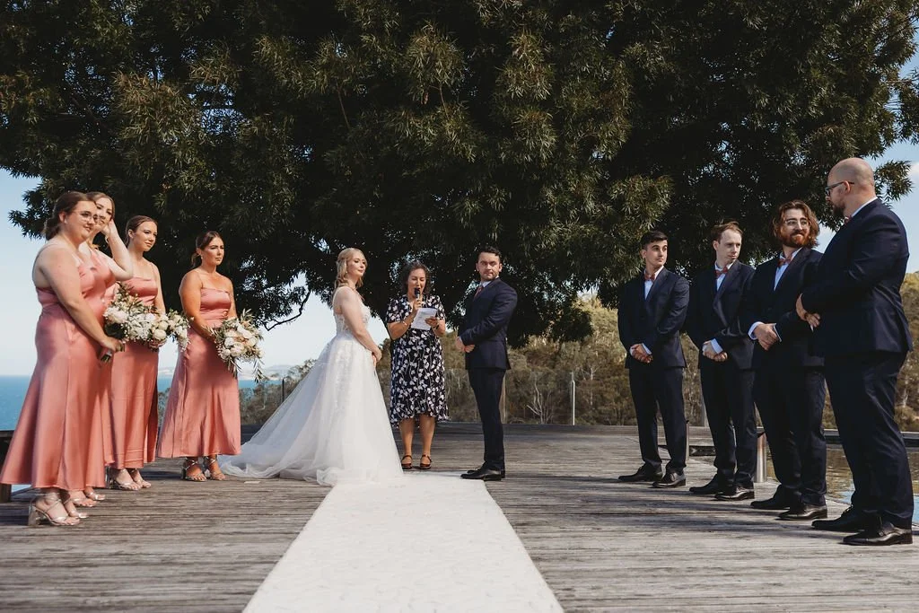 A bride and groom stand under a large tree during their outdoor wedding ceremony, with bridesmaids in pink dresses on the left and groomsmen in dark suits on the right, as a woman reads vows or vows or a speech.