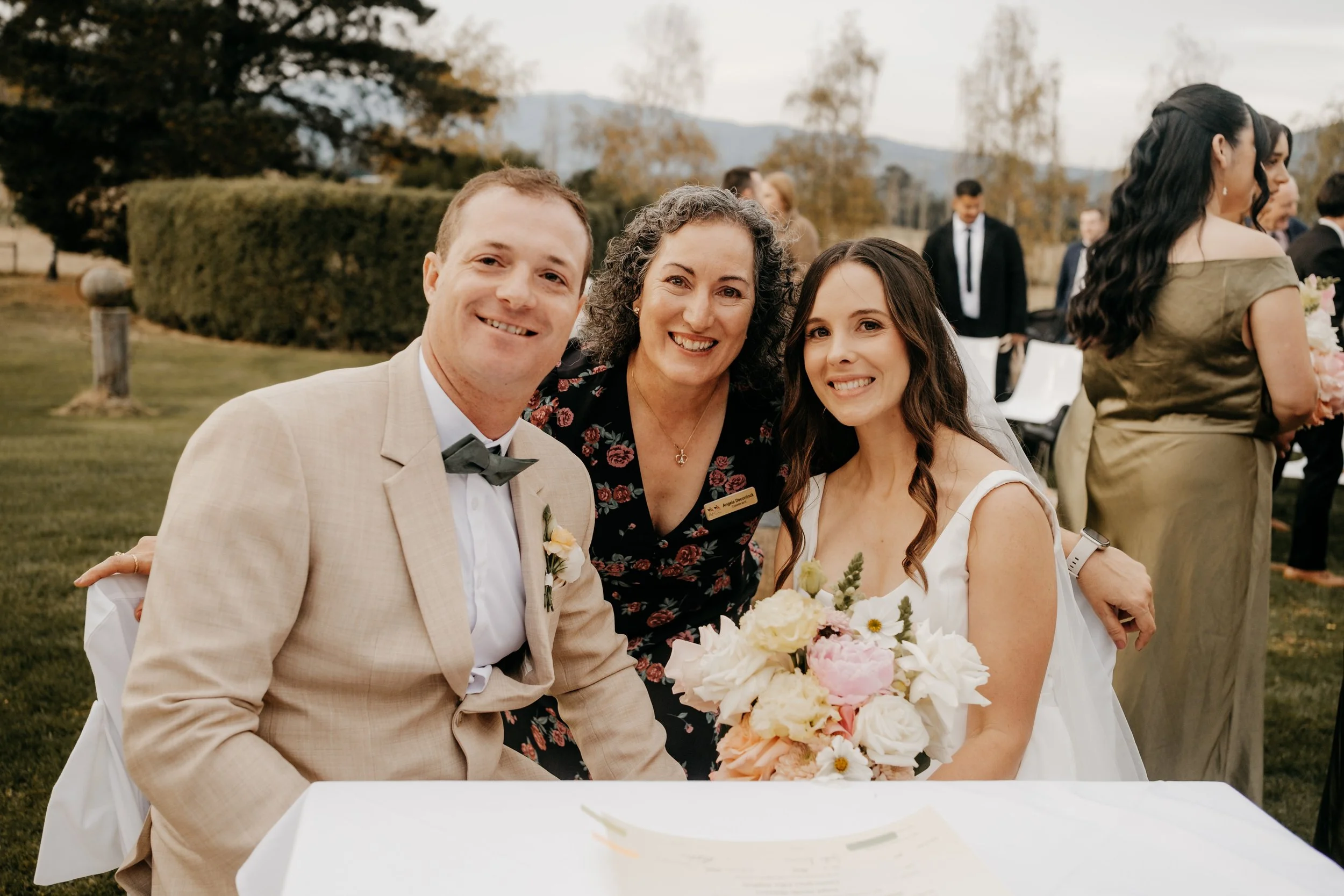 A group of three people at a wedding celebration: a man in a beige suit with a black bow tie, a woman in a white wedding dress holding a bouquet, and a woman in a black floral dress. They are outdoors in a garden with other guests and trees in the ba