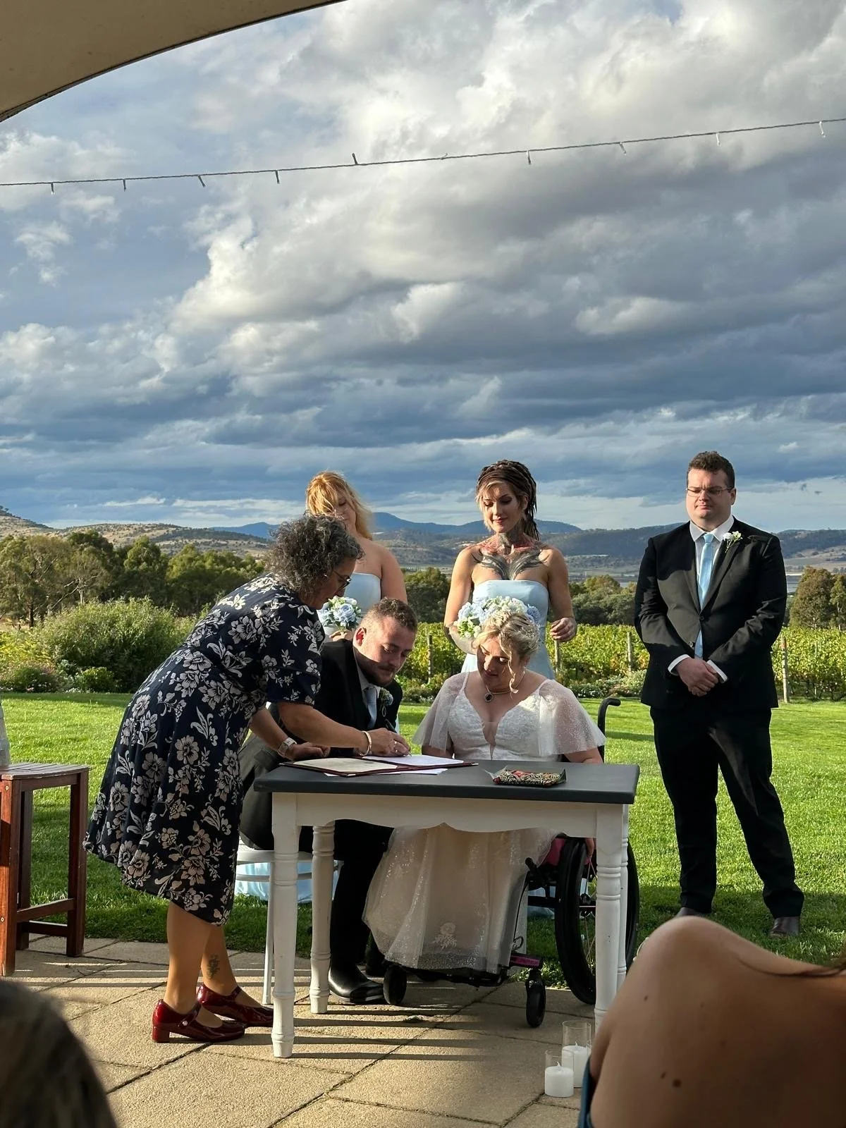 A wedding ceremony outdoors with a bride, groom, two bridesmaids, and a man in a suit standing nearby, with the officiant signing a document at a table and guests in the foreground, set against a scenic landscape with cloudy skies.
