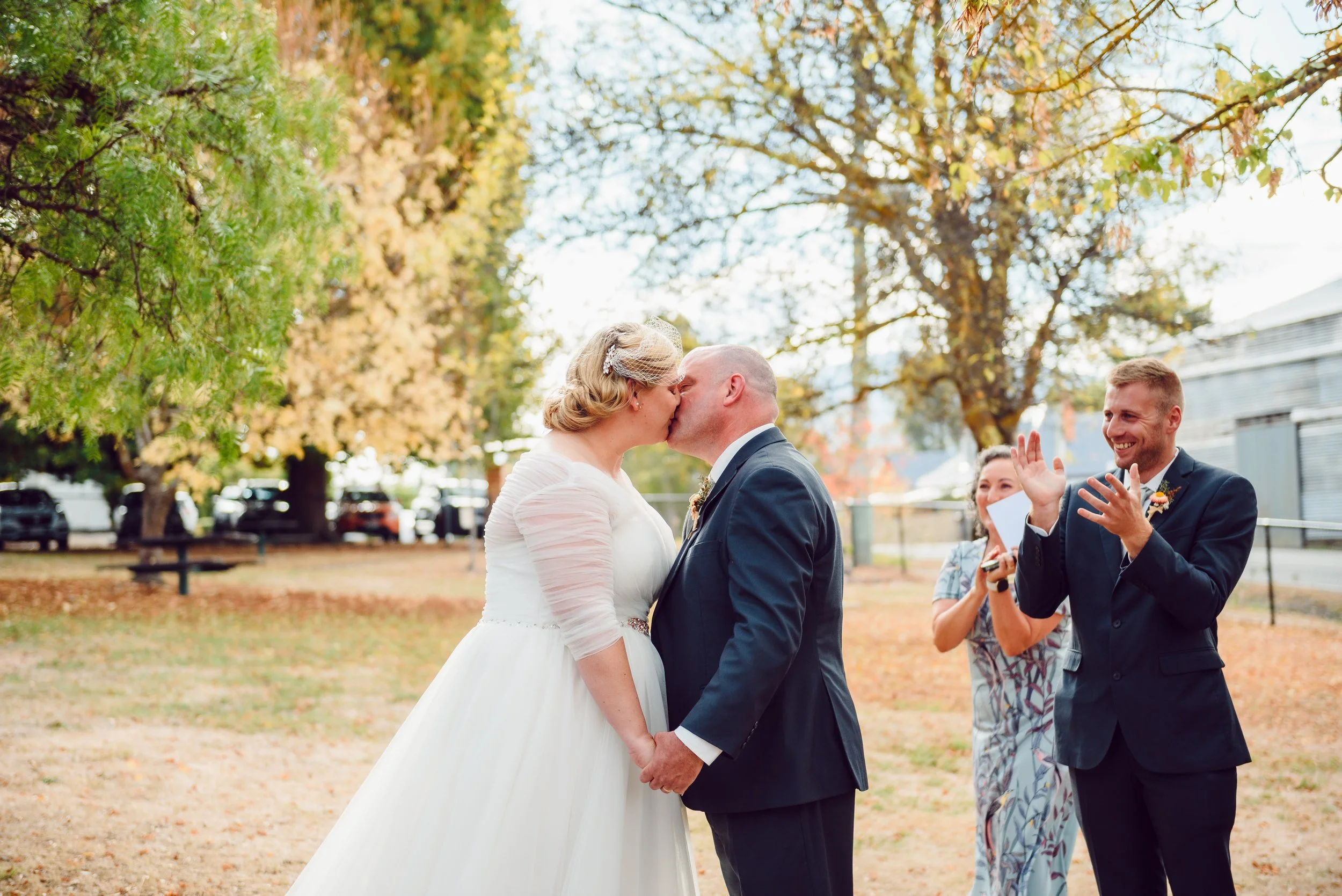 A bride and groom kiss during their outdoor wedding ceremony in fall, with a woman in a floral dress clapping and smiling in the background.