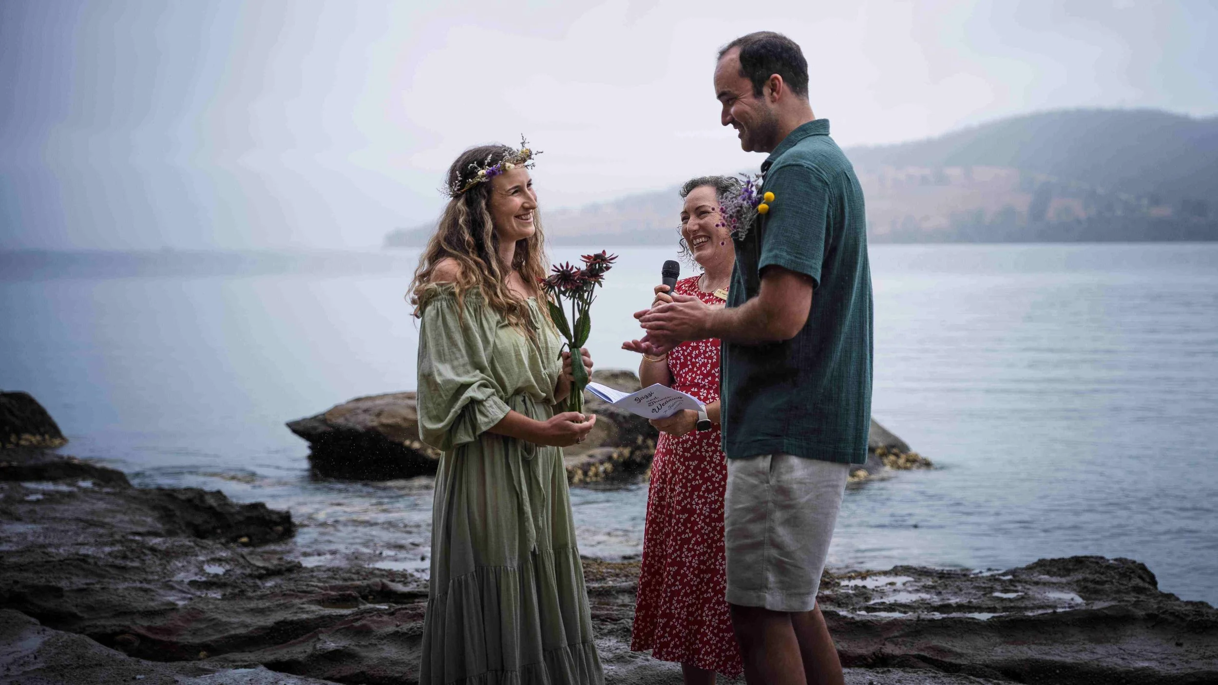 A couple is exchanging wedding vows by the sea, with a woman holding a bouquet of flowers and wearing a floral crown, and a man holding a small bouquet, while another woman officiates with a microphone and a small booklet.