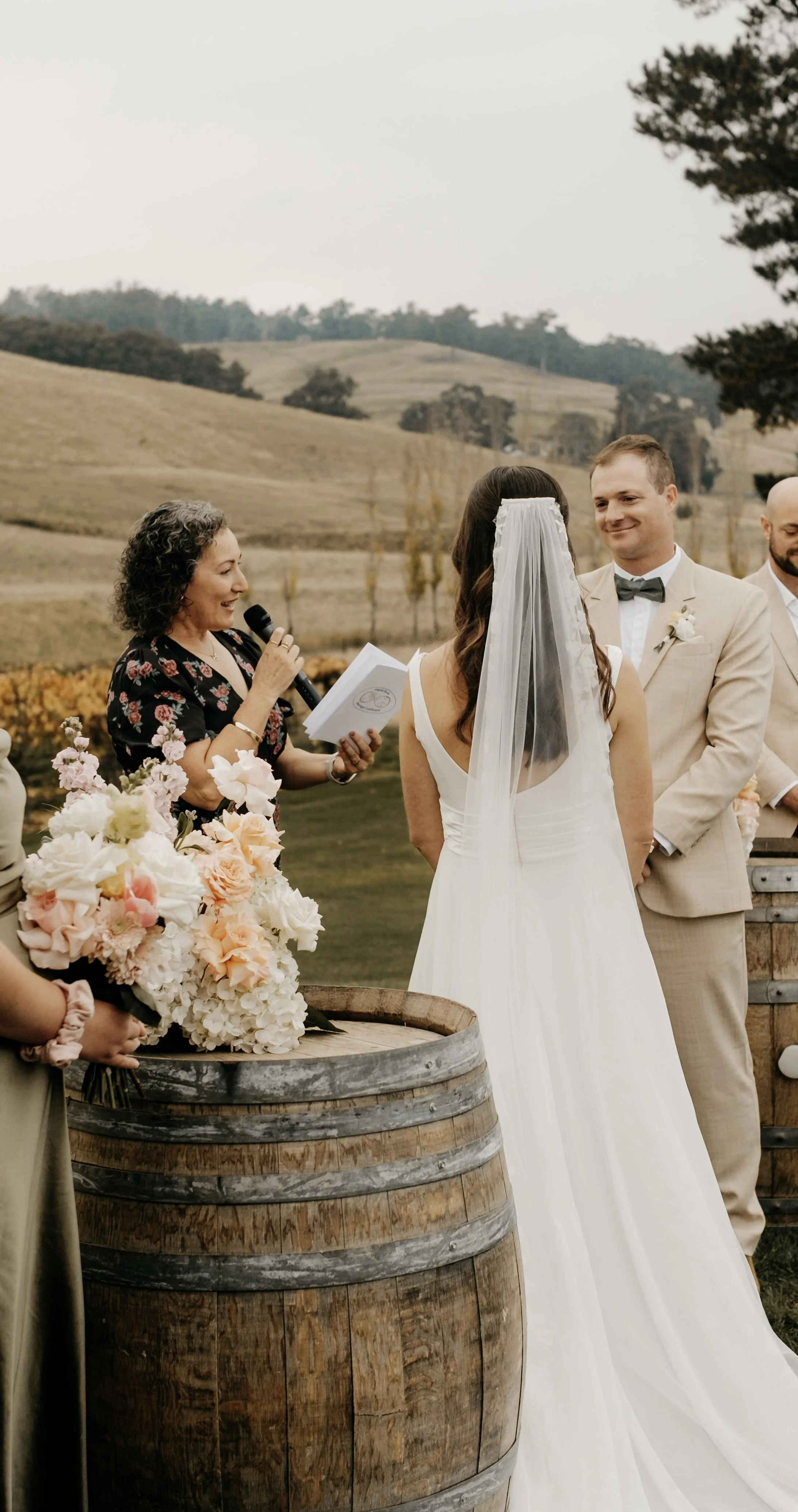 Wedding ceremony outdoors with bride, groom, officiant, and floral arrangements on a wine barrel at a beautiful Tasmanian winery.