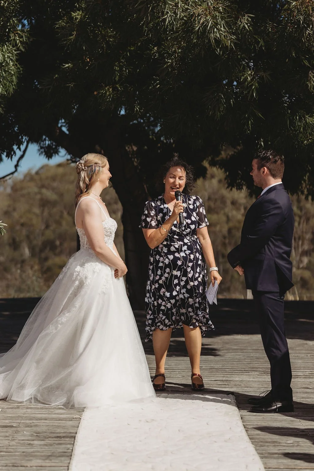 A bride and groom standing outdoors during their wedding ceremony, with a woman officiant speaking into a microphone.