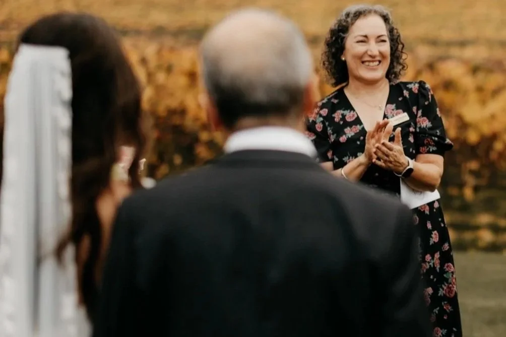 A wedding ceremony outdoors in a vineyard (Tasmanian HomeHill Winery), with a marriage celebrant smiling and clapping in front of a married couple, with autumn foliage in the background.