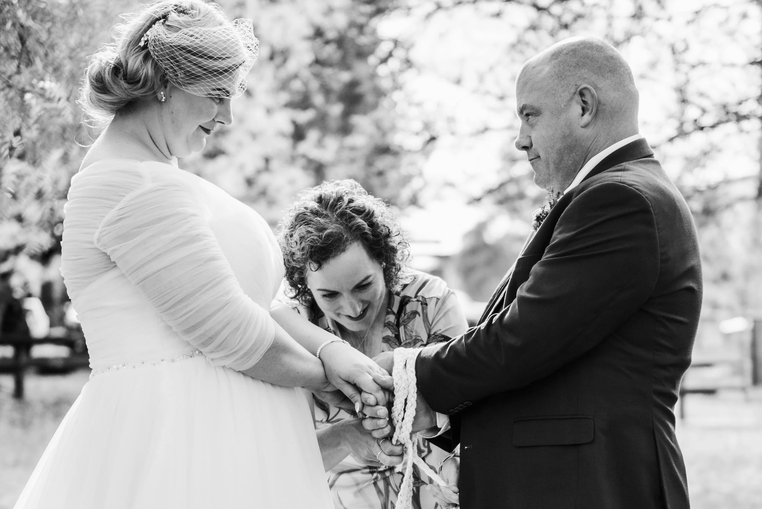 A black and white photo of a wedding ceremony outdoors, with a bride and groom holding hands and looking at each other, as an officiant or family member holds their hands.