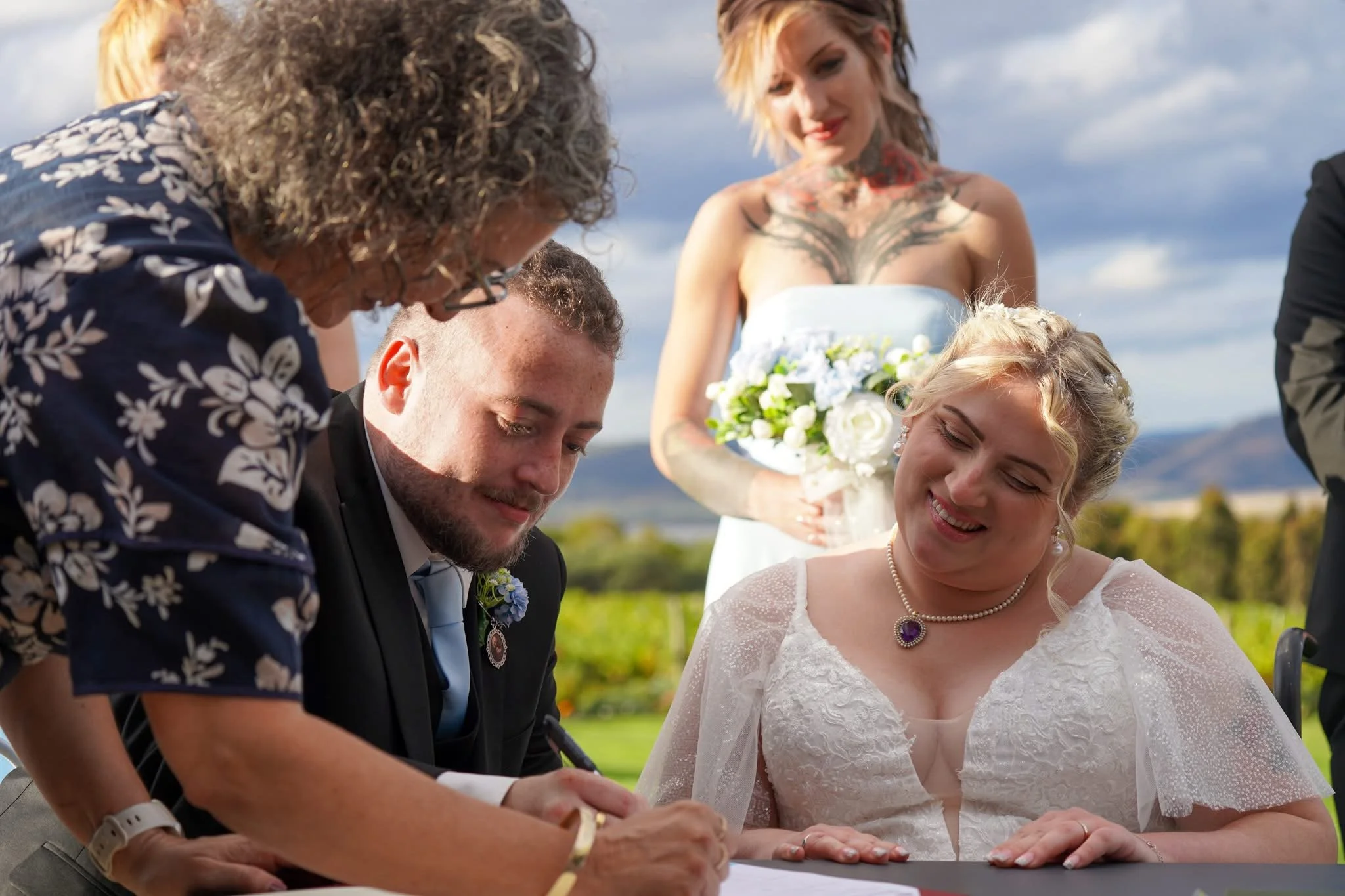 A bride and groom signing wedding documents outdoors with bridesmaids in a floral dress and a white dress with pearls happily watching.