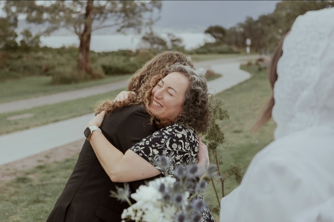 Groom hugging his marriage celebrant in gratitude while being fondly looked upon by beautiful bride in a garden outdoors on a grassy area near a path, with trees and water in the background, during daytime.