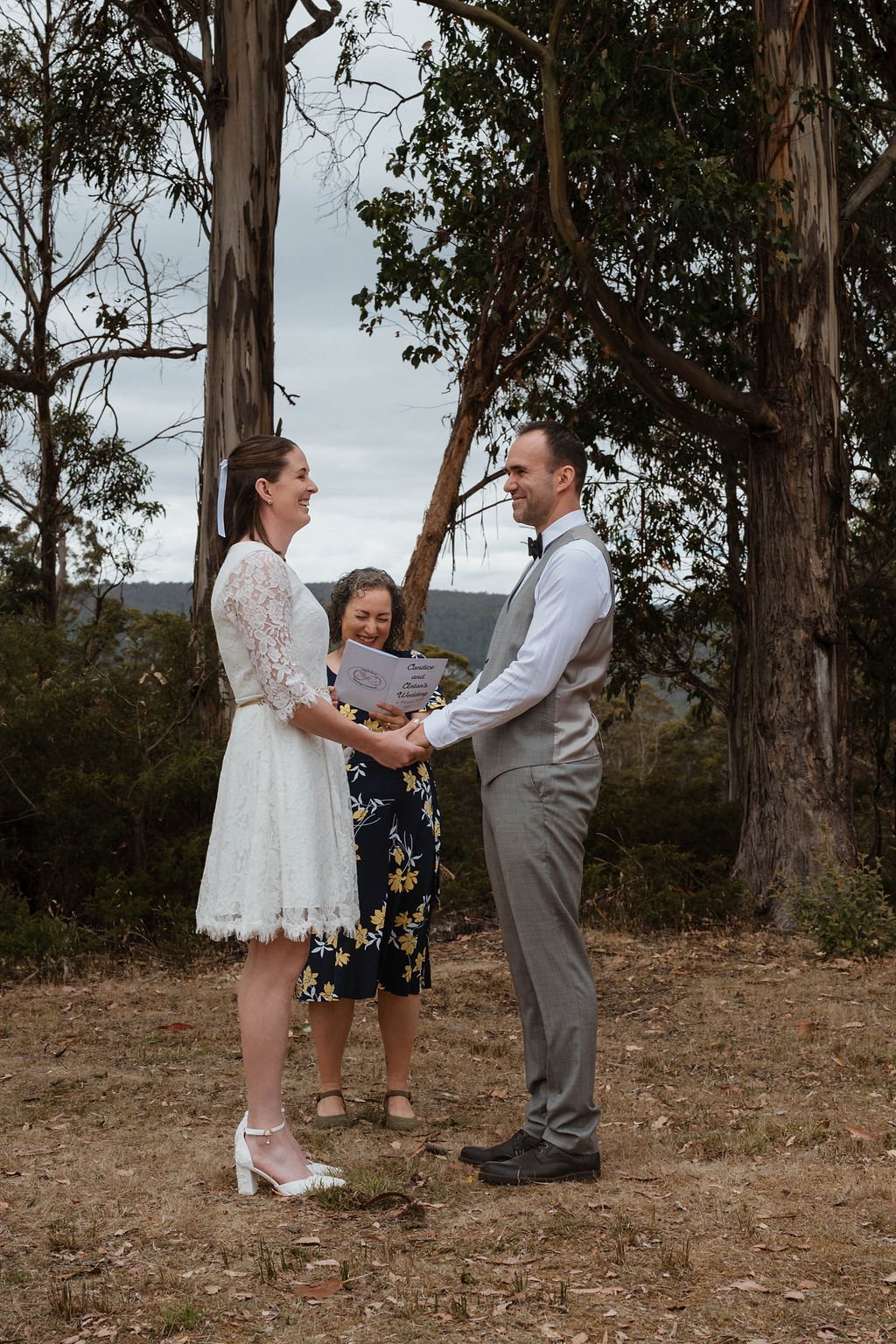 A couple exchanging vows outdoors, holding hands and smiling, during a wedding ceremony with an officiant standing behind them, set in a wooded area with tall trees and a cloudy sky.