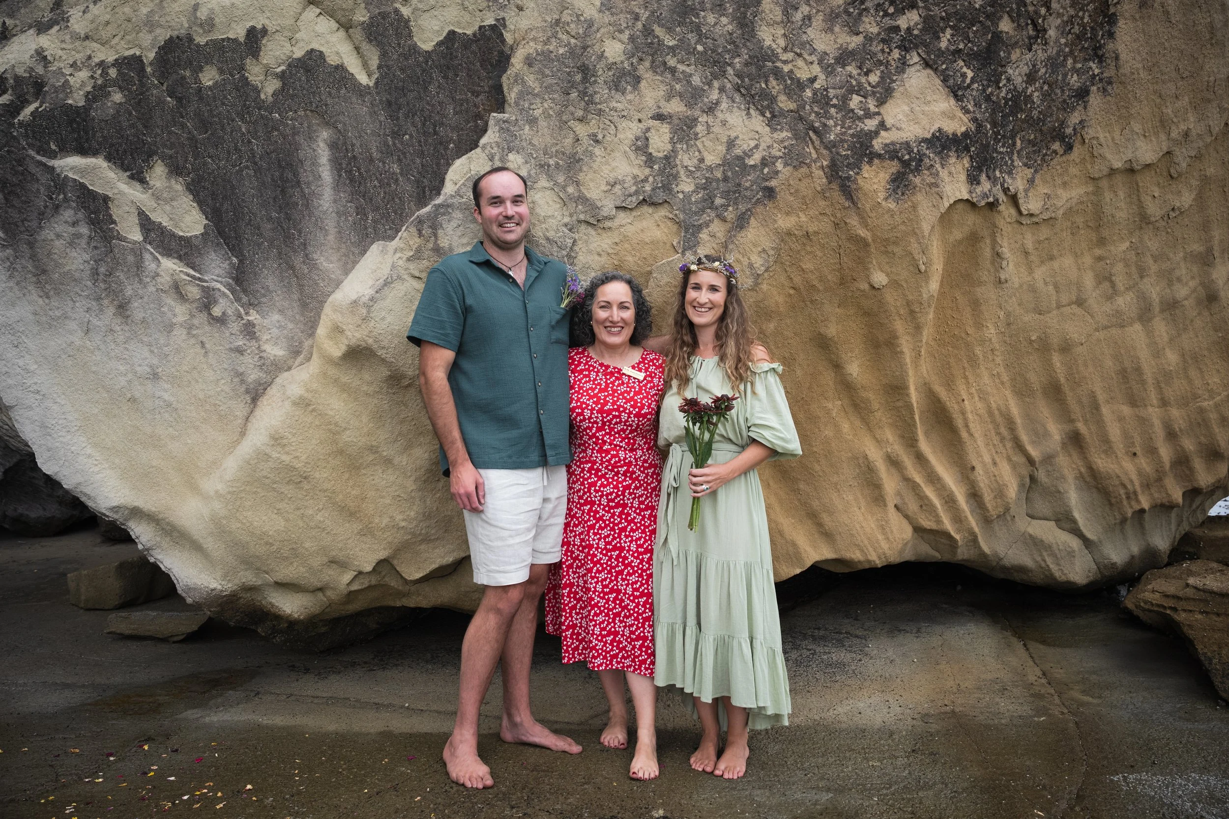 Three people standing barefoot on a beach in front of a large rock formation. The person on the left is a man wearing a green shirt and white shorts, smiling. The woman in the center is wearing a red dress with white patterns and a flower crown, hold