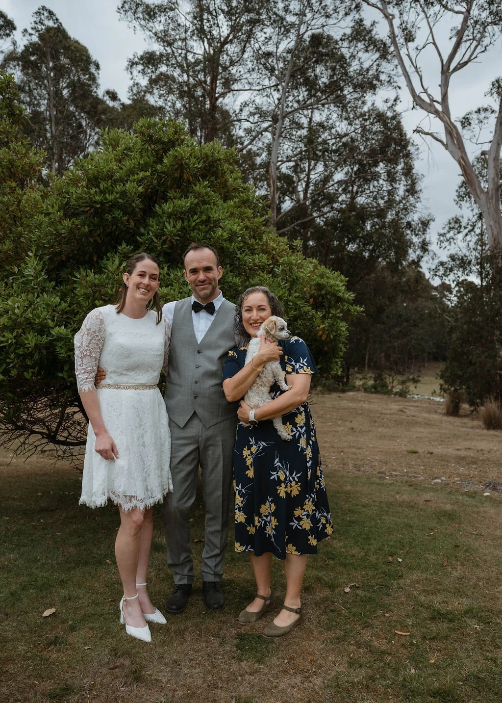 A group of three people, two women and a man, outdoors in a natural setting with trees and bushes in the background. The woman on the left wears a white lace dress and white heels, the man in the middle wears a gray vest and bow tie, and the woman on