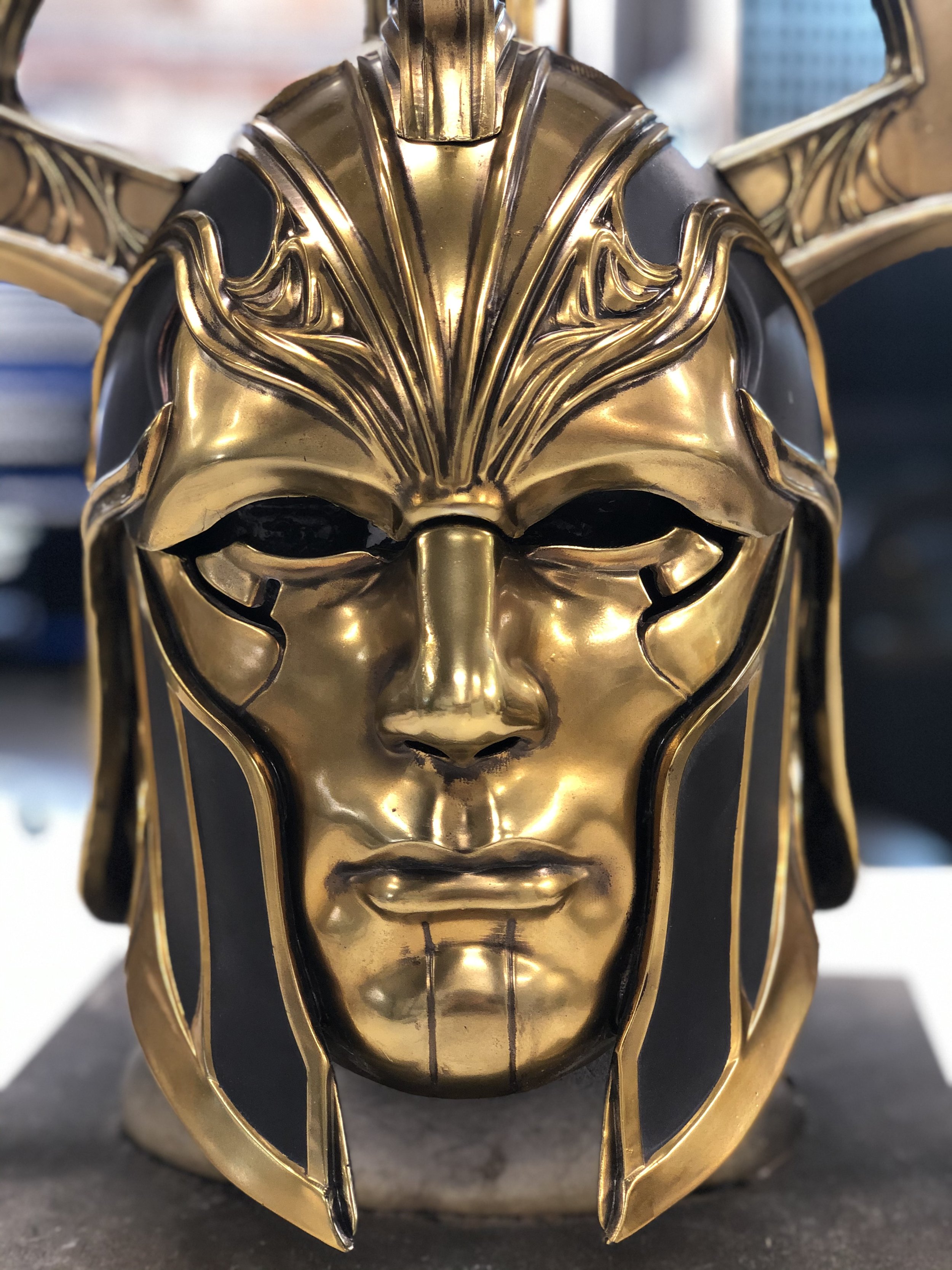 Close-up of a decorative gold and black mask resembling a regal or mythological face with intricate details and a stern expression.