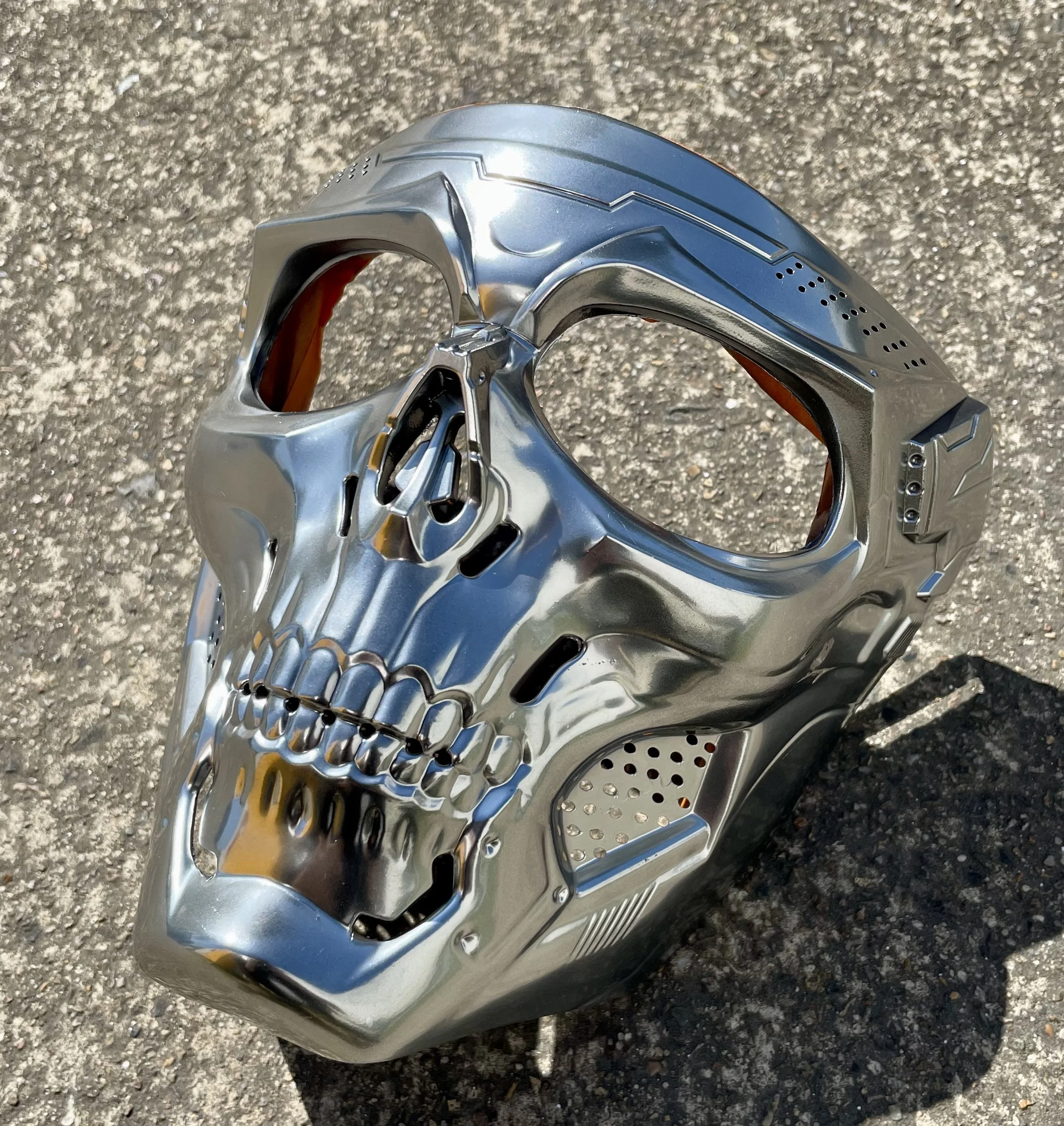 A metallic skull mask with futuristic design, placed on an asphalt surface.