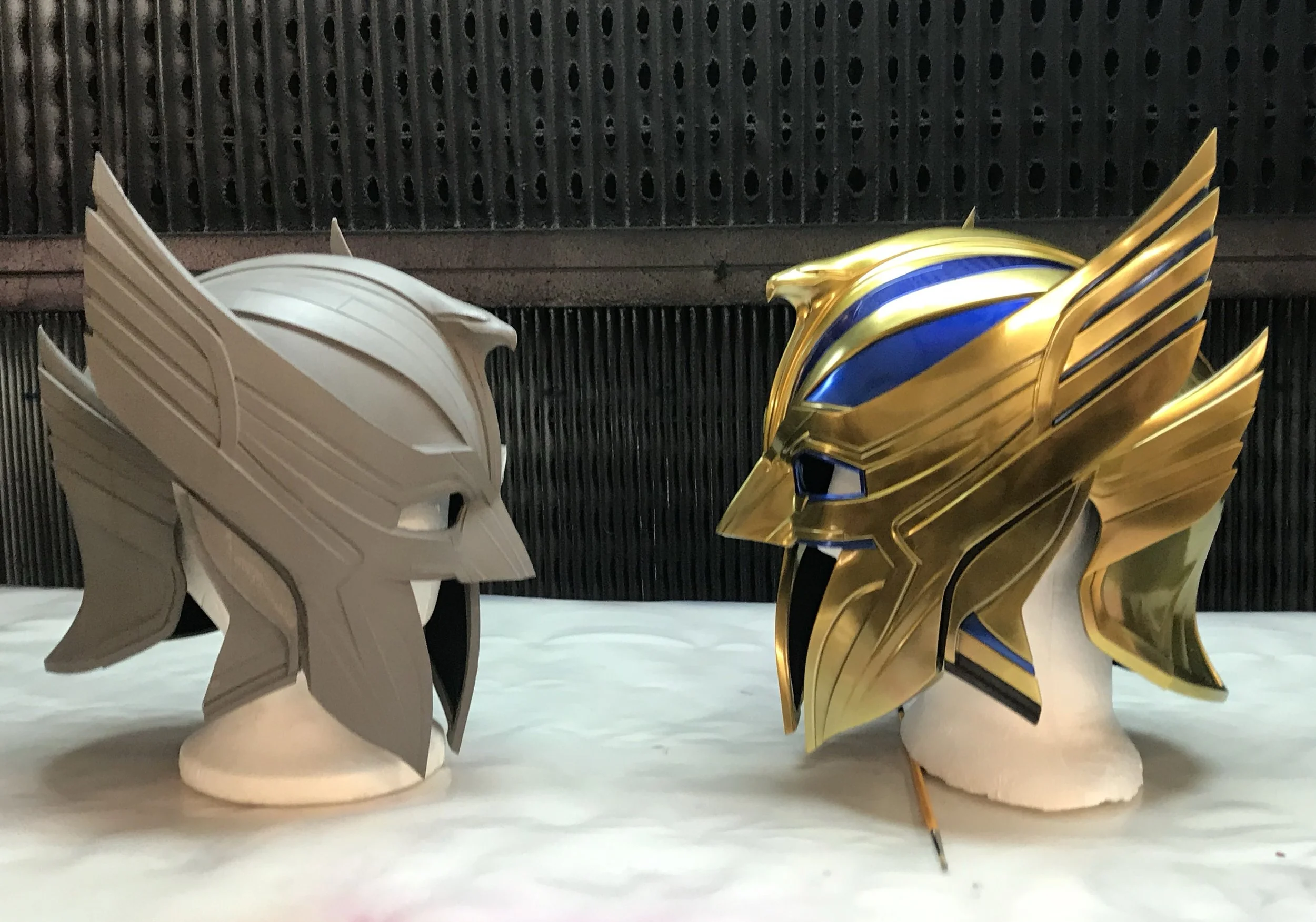 Two detailed helmet models inspired by the character Genji from Overwatch are displayed on a table, one in matte silver and the other in shiny gold with blue accents.