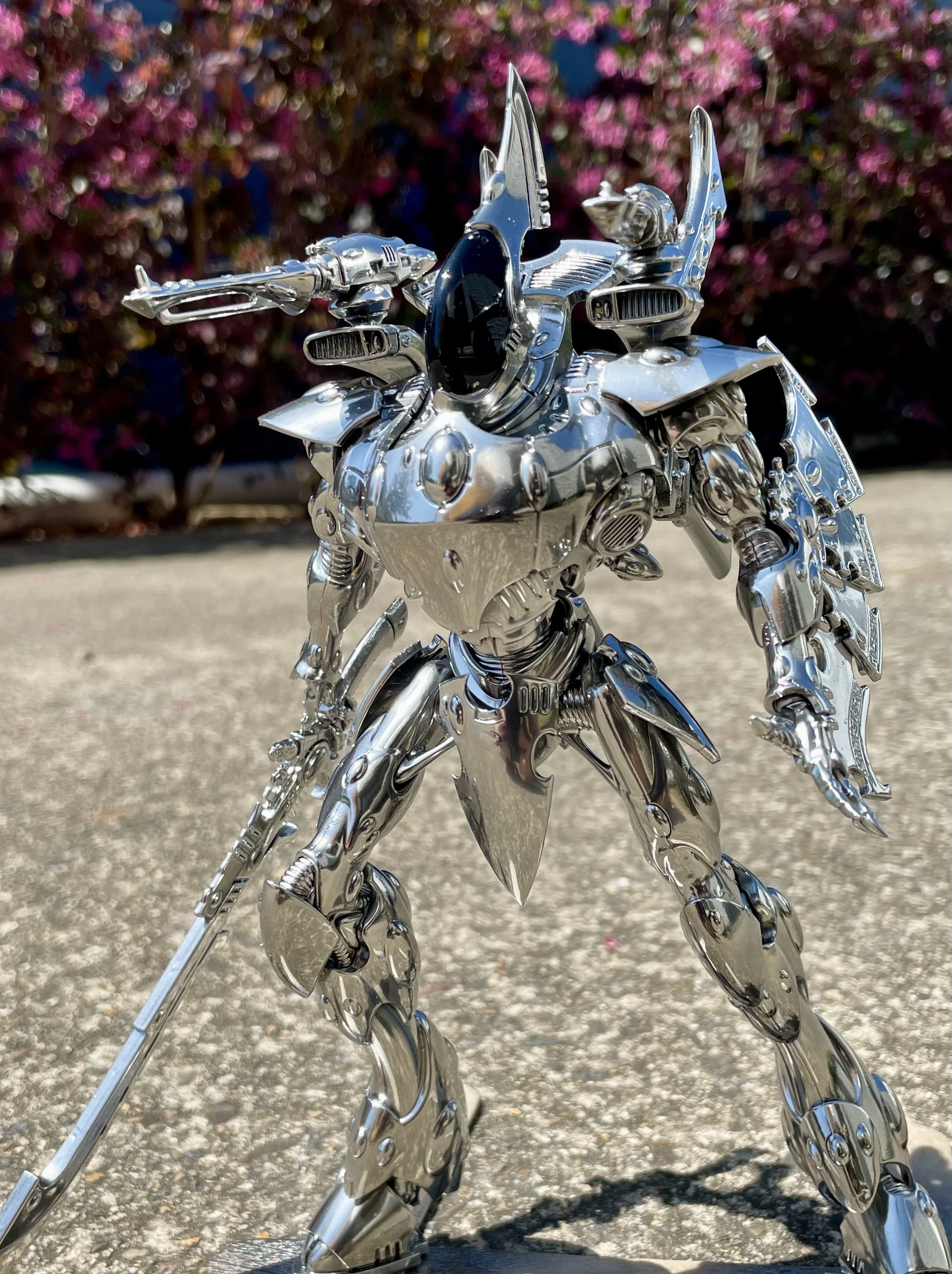 A detailed metallic robot figure with a futuristic design, posed outdoors on a concrete surface with pink flowering bushes in the background.