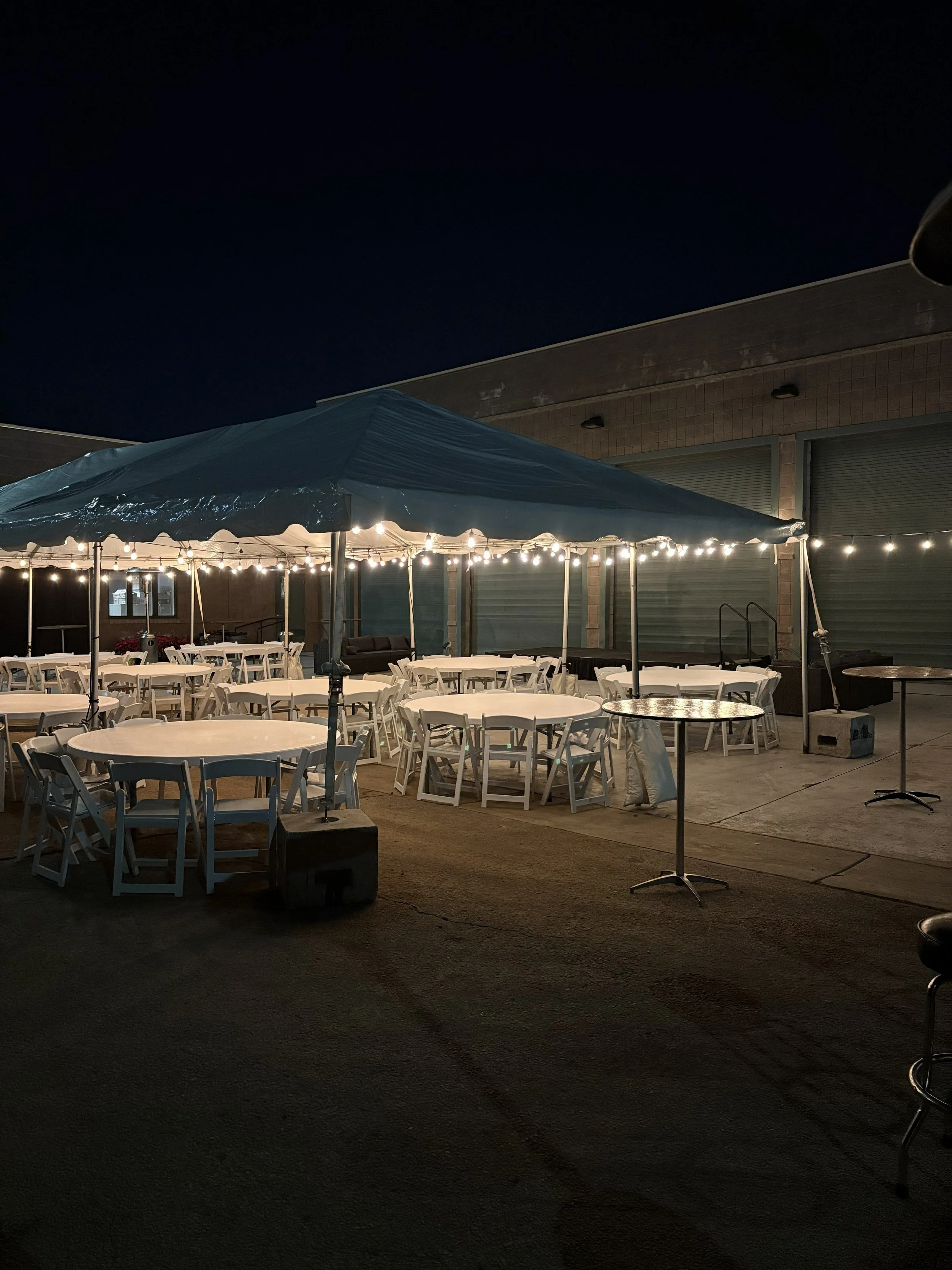Outdoor event setup with white round tables and chairs under a large canopy tent decorated with string lights, at night.
