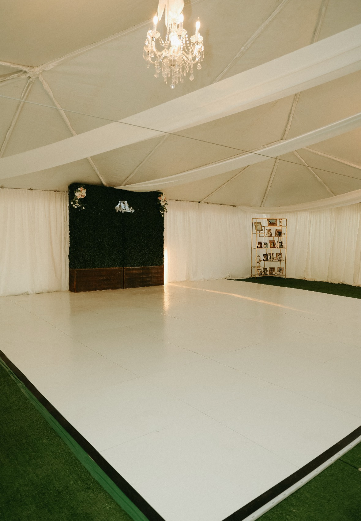 Event space with a white dance floor, elegant drapes, a chandelier, a black decorative wall with floral accents, and a photo display shelf in the corner.