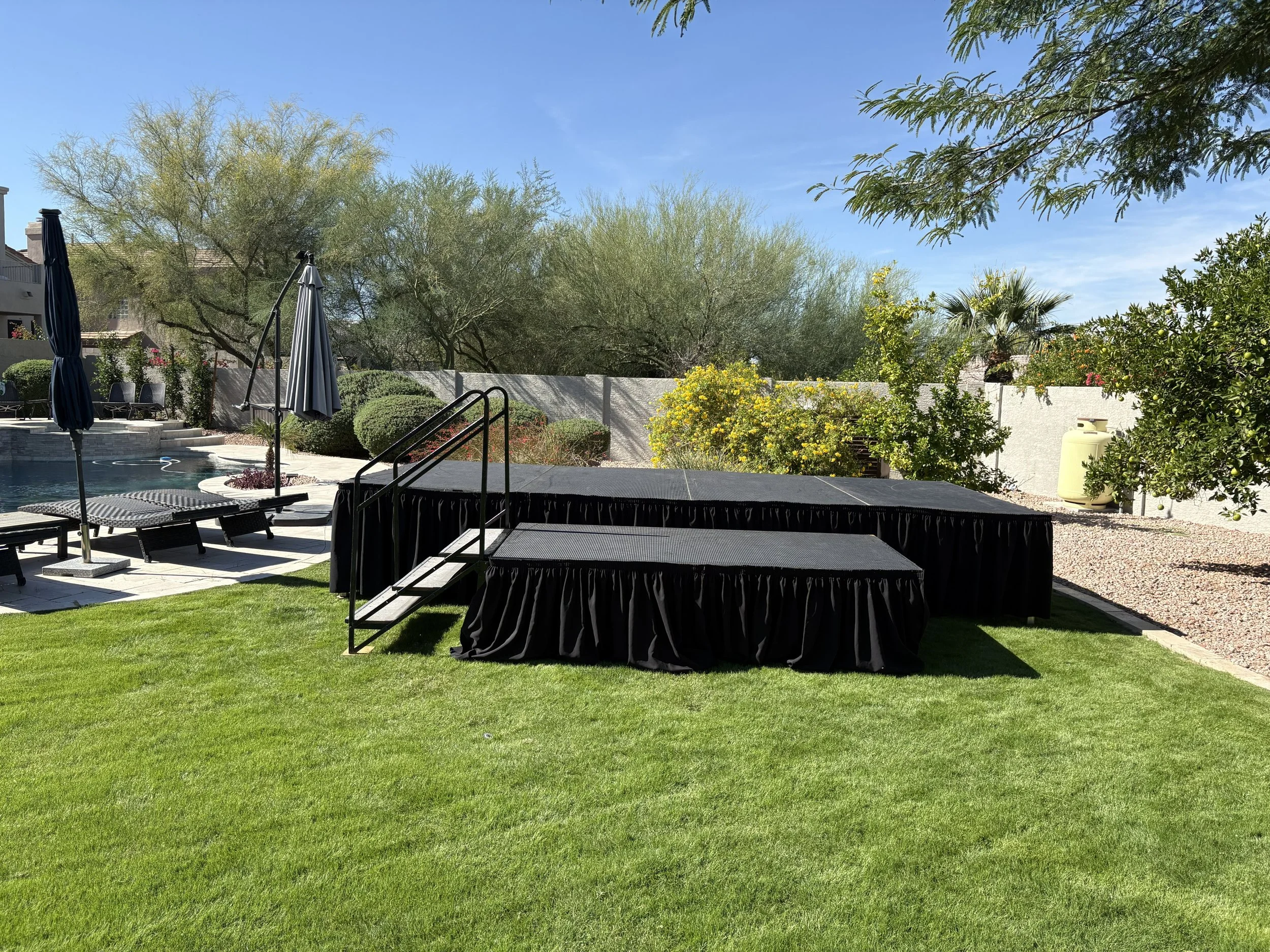 An outdoor backyard scene with a swimming pool, lounge chairs, umbrellas, and a stage with black drapes set on green grass, surrounded by trees and a white fence.
