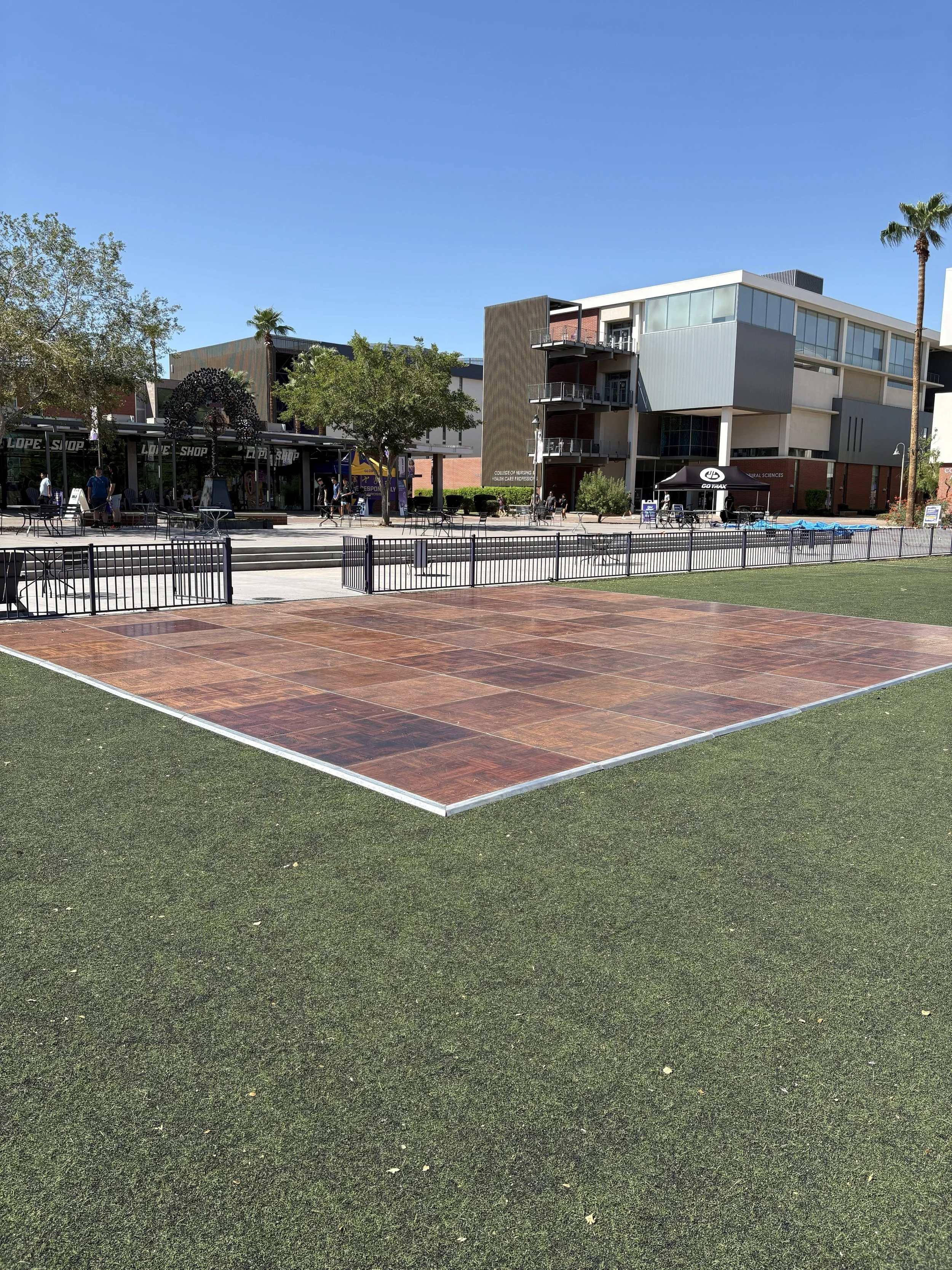 A public outdoor space with a wooden dance floor area, surrounded by artificial grass, in front of a modern building with shops and people walking.