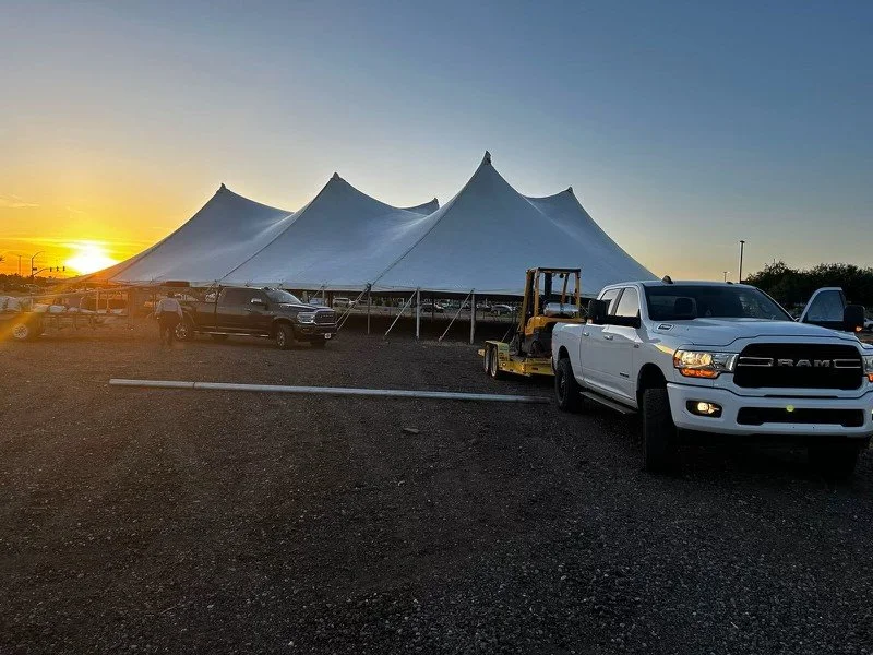 Large white tent with peaked tops, set up in an open area during sunset. Several pickup trucks and a forklift are parked in front of the tent on a gravel surface.