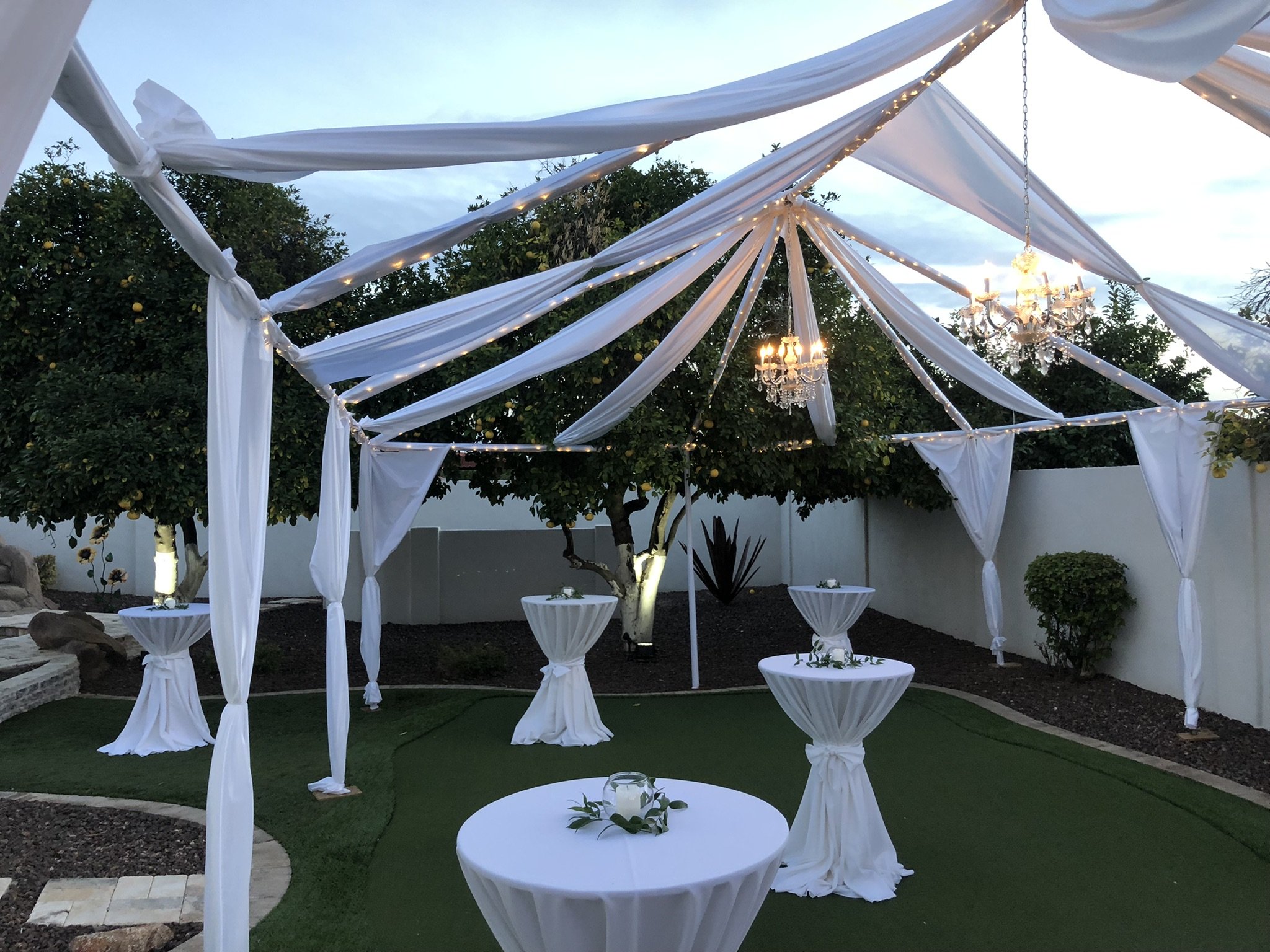 Outdoor event setup with white draped fabric canopy, chandeliers hanging from the structure, small round tables with white cloths and small floral decorations, green artificial grass, trees with yellow fruits, and a white wall in the background.