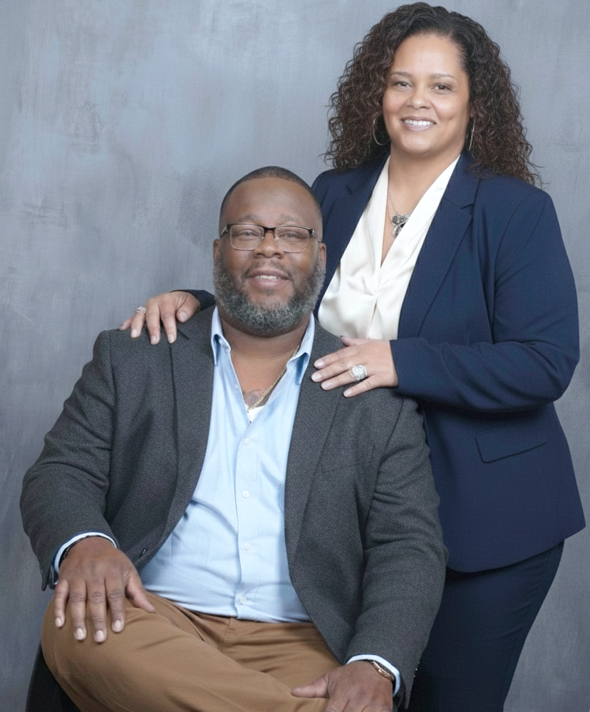 A woman with curly hair and a man with glasses and a beard pose together against a gray background. The woman stands behind, resting her hand on the man's shoulder, while the man sits with his legs crossed.