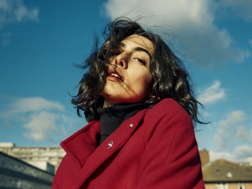 A woman with dark, curly hair wearing a red coat and a black turtleneck, outdoors against a blue sky with some clouds.