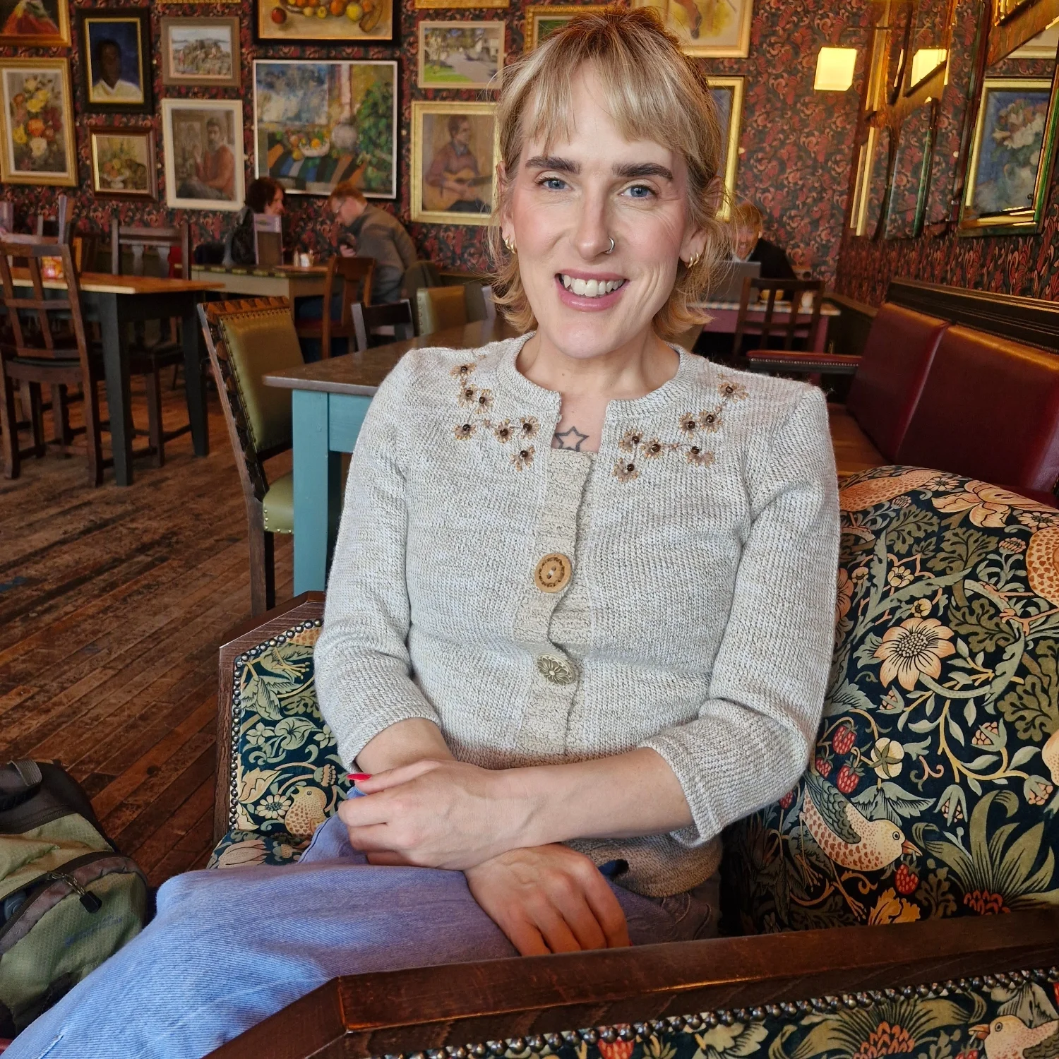 A woman with short blonde hair and a nose piercing, wearing a beige cardigan with floral embroidery, seated in a cosy coffeehouse or restaurant with framed artwork on the walls.